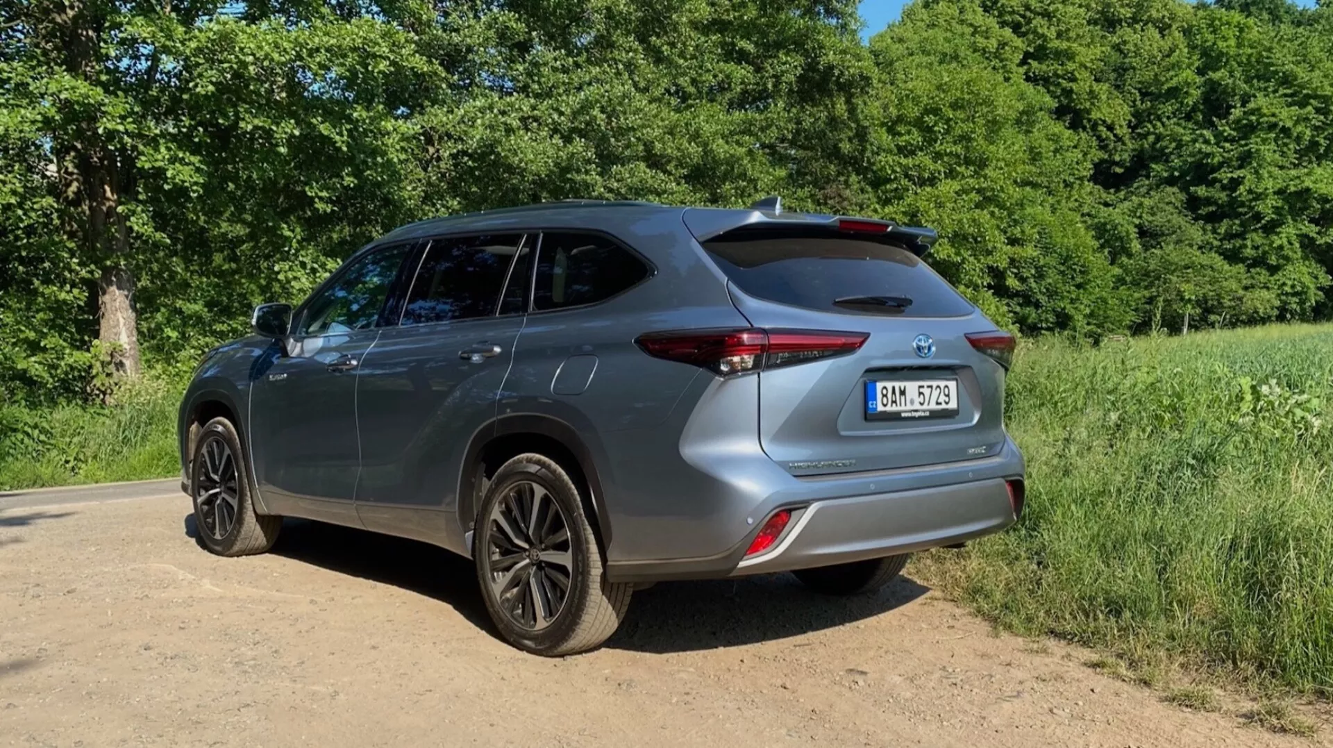 Toyota Highlander 2021 Hybrid Business Edition ukazany z tylnego trójkwartalowego widoku, podkreślający prawą stronę i tył samochodu. Ujęcie na poziomie gruntu eksponuje ciemne felgi i całkowitą wysokość pojazdu. Tylny design, z eleganckimi światłami, rzeźbionym zderzakiem i tylną klapą ze spojlerem, jest zaakcentowany. Obraz również pokazuje design drzwi, bocznych okien i lusterek. Pojazd jest umieszczony na gruntowej lub żwirowej ścieżce z zieloną roślinnością i drzewami w tle. Słoneczne oświetlenie rzuca cienie do przodu, sugerując, że słońce znajduje się za kamerą.
