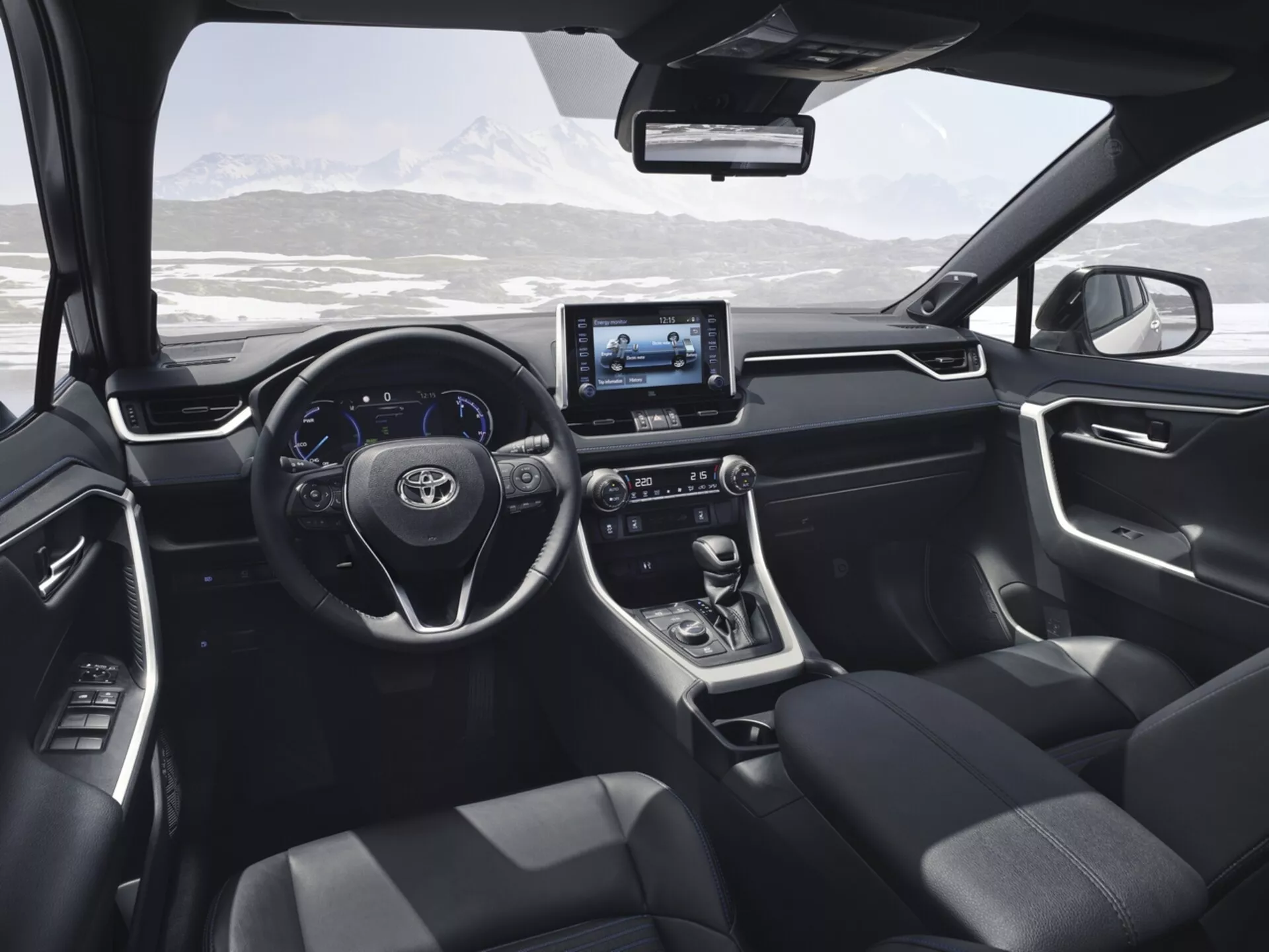 Widok wnętrza Toyota RAV4 2019 z tylnego siedzenia, prezentujący nowoczesny design i funkcje technologiczne, w tym kierownicę z przyciskami sterowania, cyfrowy wyświetlacz, dotykowy system infotainment, dźwignię zmiany biegów automatycznej skrzyni biegów, ciemno tapicerowane fotele oraz panele sterowania na drzwiach z widokiem na malowniczy, zaśnieżony krajobraz górski przez przednią szybę.