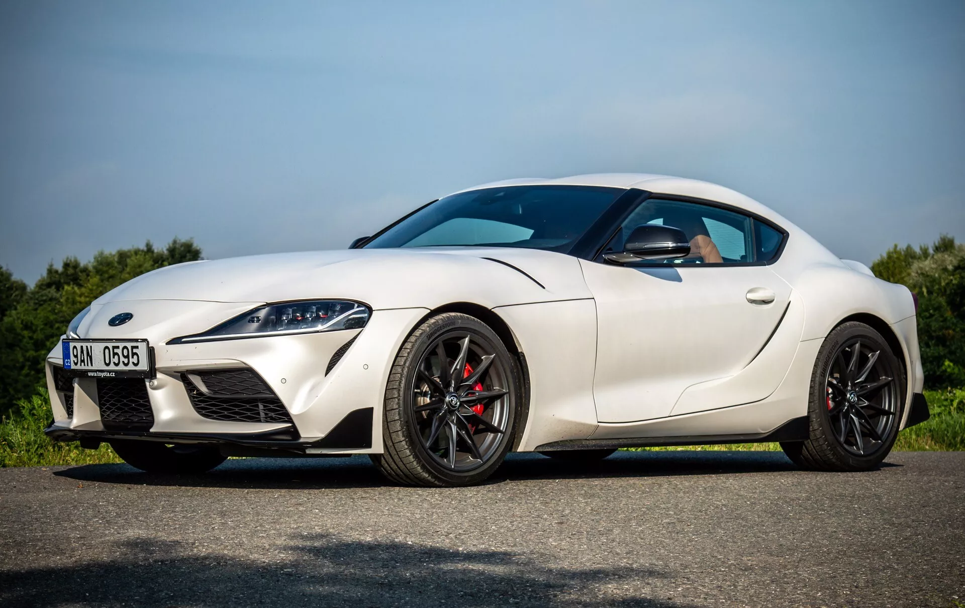 Biały sportowy coupe Toyota Supra (GR-Supra) z 2019 roku zaparkowane na asfalcie pod jasnym niebem, widok z boku pokazujący reflektory LED, sportowy aerodynamiczny design i cechy zorientowane na osiągi