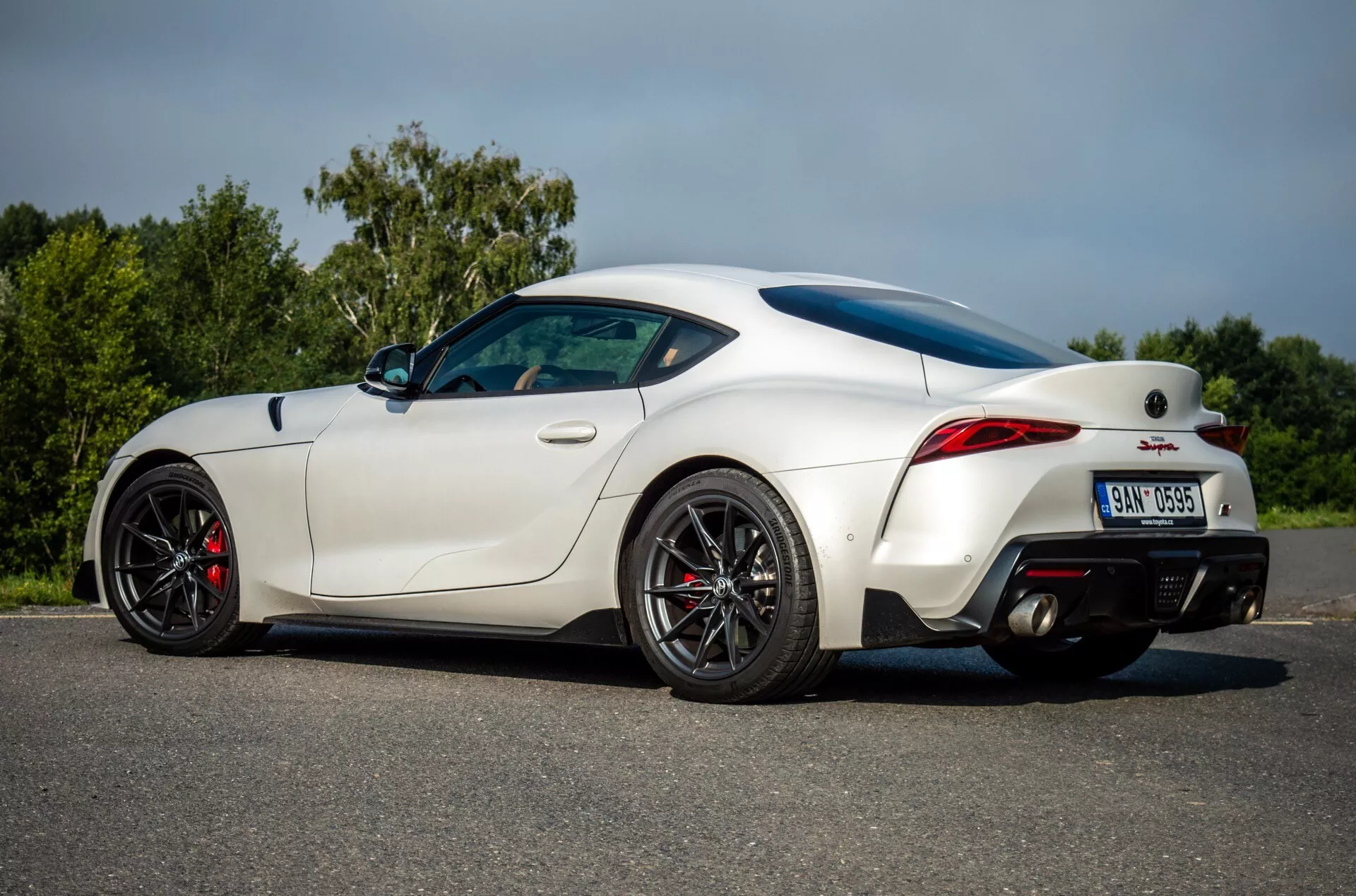 Toyota Supra (GR-Supra) z 2019 roku w kolorze białym prezentująca swój aerodynamiczny design, sportową sylwetkę ze spoilerem tylnym, wysokowydajne czarne koła z czerwonymi zaciskami hamulcowymi, podwójny system wydechowy oraz nowoczesne tylne światła LED z perspektywy boczno-tylnej