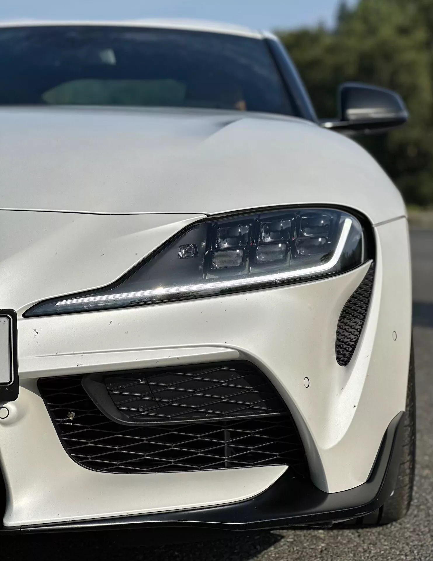 Zbliżony, kątowy widok z przodu-lewej strony Toyoty Supra (GR-Supra) z 2019 roku prezentujący nowoczesne światło LED, przedni zderzak o orientacji sportowej z wlotem powietrza w siatce o strukturze plastra miodu, aerodynamiczne kontury i smukłe lusterko boczne na błyszczącym wykończeniu