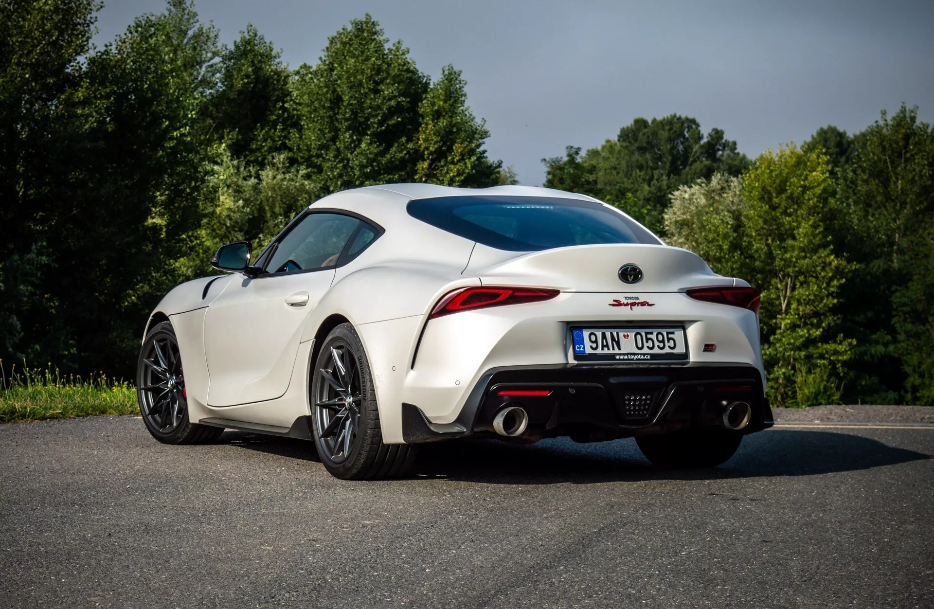 GR-Supra 2019 w kolorze białym, widok z tyłu z boku ukazujący sportowy design z zakrzywionymi tylnymi bokami, charakterystycznymi felgami, lusterkami bocznymi, tylnymi światłami LED, podwójnymi wyjściami wydechowymi i zintegrowanym spojlerem, otoczony zielenią.