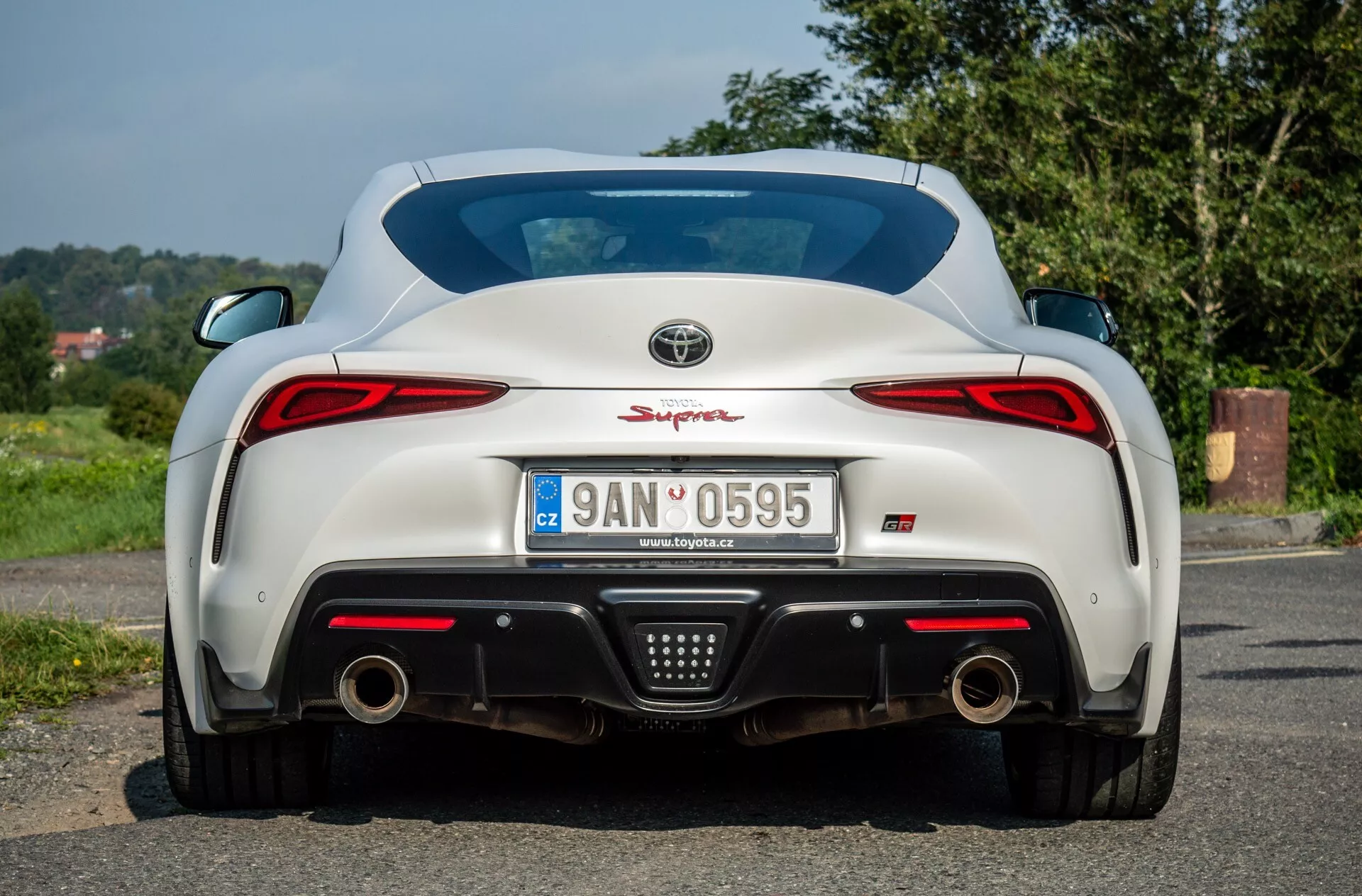 Tylny widok Toyoty Supra (GR Supra) z 2019 roku prezentujący światła tylne LED, podwójny układ wydechowy, dyfuzor tylny, czujniki parkowania i zintegrowany spoiler na błyszczącej białej powierzchni, zaparkowany na asfalcie z naturalnym, wiejskim tłem