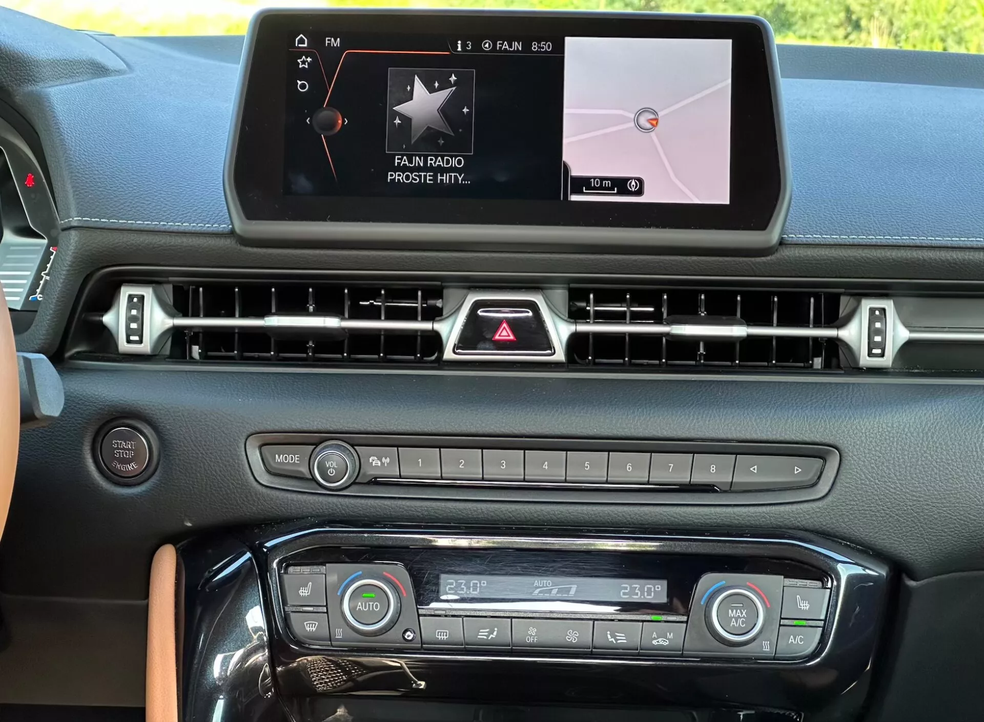 Widok wnętrza kokpitu Toyota Supra GR 2019 ukazujący konsolę środkową z cyfrowym ekranem dotykowym systemu infotainment wyświetlającym 'FAN RADIO' i nawigację mapy, rząd nawiewów, przycisk świateł awaryjnych, przyciski i pokrętła sterowania mediami i klimatyzacją z ustawieniem temperatury podwójnej strefy na 23°C, oraz elementy designu z kontrastowym wykończeniem.