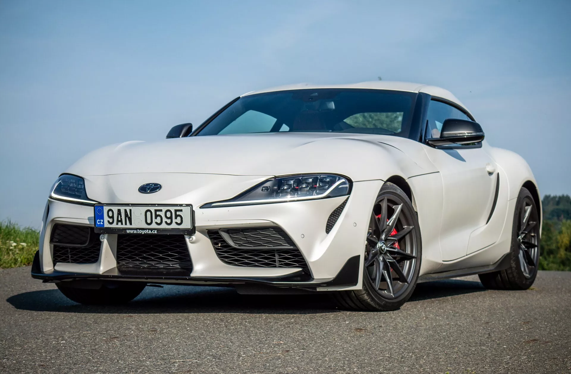 Biały Toyota Supra GR 2019 widziany z niskiego przedniego kąta trzy czwarte podkreślający jego aerodynamiczny profil, wąskie reflektory LED, duże wloty powietrza, splitter przedni i sportowo ukształtowaną maskę.