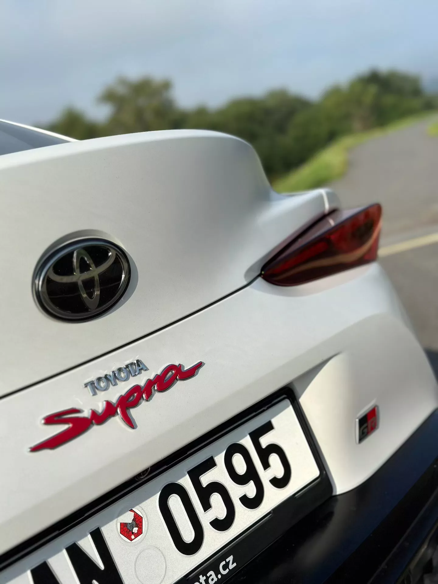Zbliżenie na tylną część białej Toyoty Supra z 2019 roku, przedstawiające emblem Toyota, czerwony napis modelu 'Supra', prawe tylne światło LED, wgłębioną przestrzeń na tablicę rejestracyjną, część tylnego zderzaka, zintegrowany spojler, część dachu i tylną szybę, na tle zamazanej drogi i zieleni.