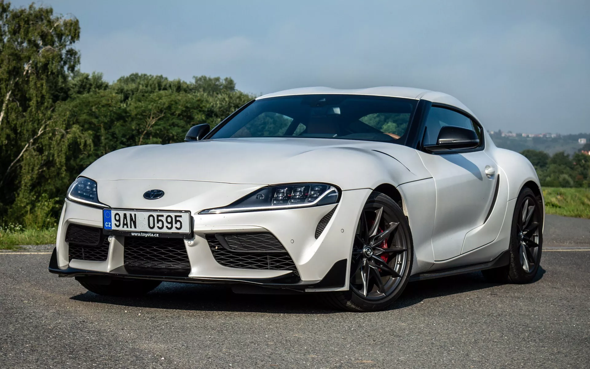 Sportowy samochód Toyota GR Supra 2019 z przodu ujęcie trzy czwarte ukazujące aerodynamiczną konstrukcję, elegancką przednią kratkę, czarne felgi aluminiowe o wielu szprychach i błyszczący biały lakier na asfalcie z zielonym tłem