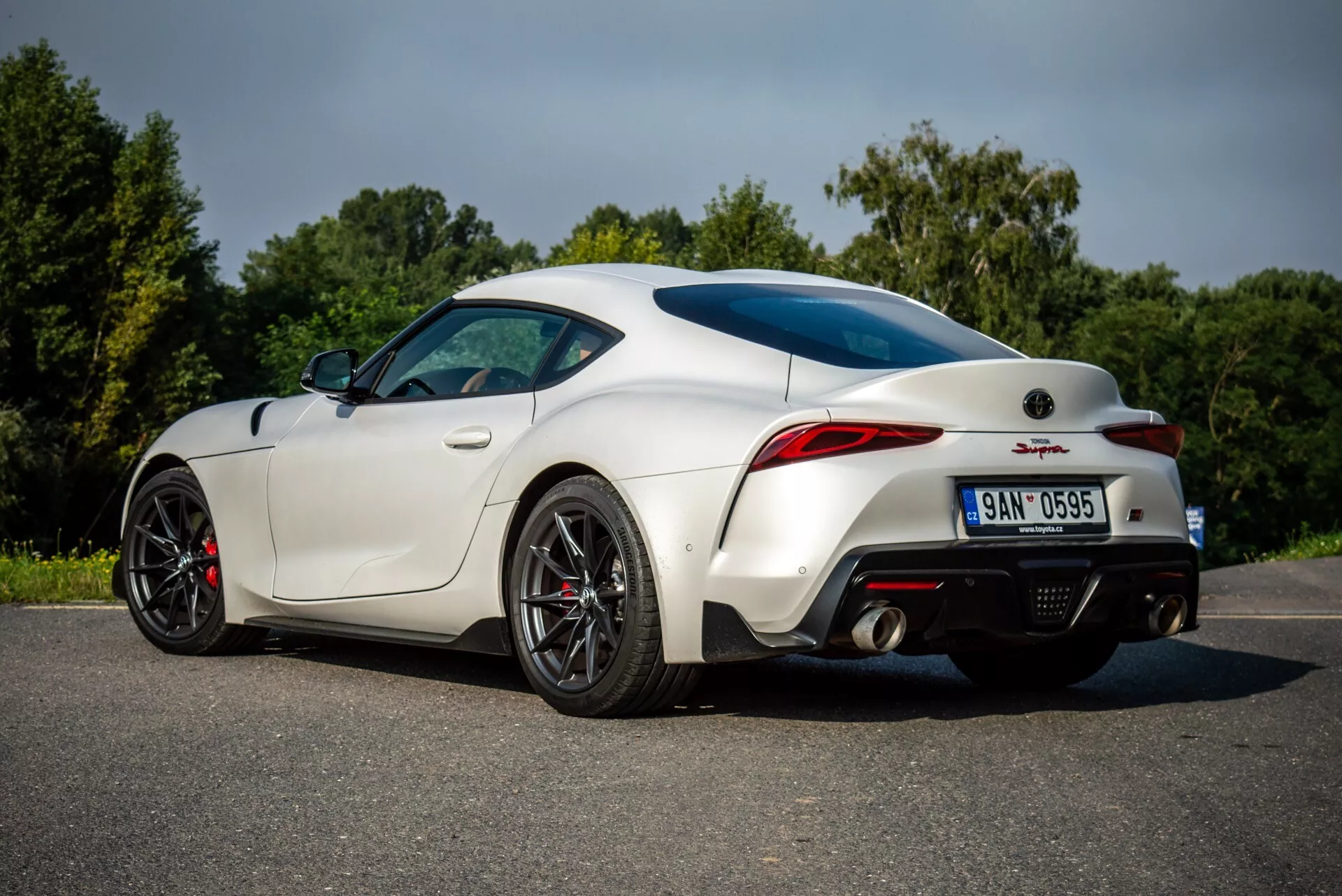 Toyota Supra GR 2019 w kolorze białym, prezentująca swój aerodynamiczny design z widoku trzy czwarte od tyłu i boku pasażera na asfalcie, łącznie z charakterystycznymi tylnymi światłami LED, kołami o wysokiej wydajności z czerwonymi zaciskami hamulcowymi, nowoczesnym systemem wydechowym i tylnym dyfuzorem poprawiającym aerodynamikę.