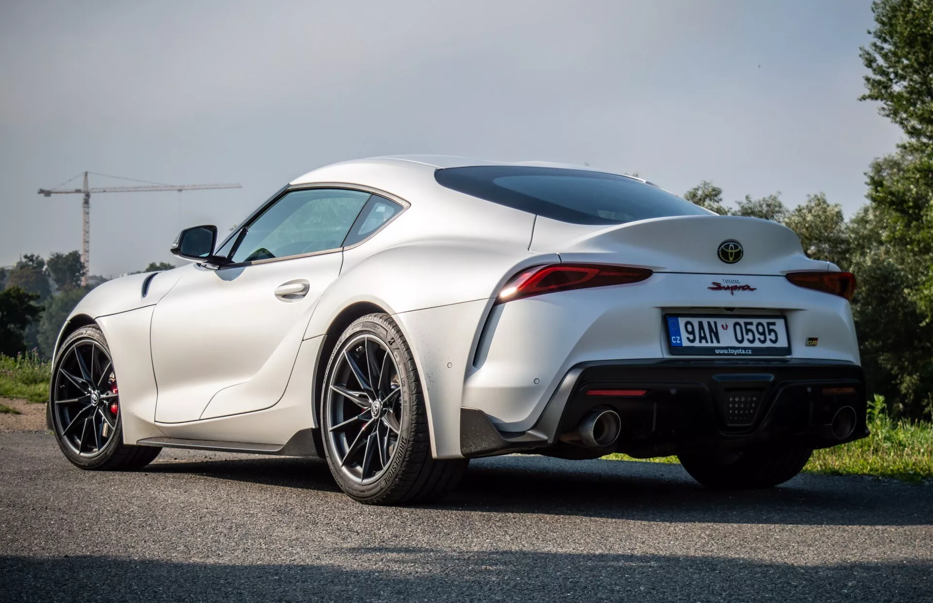 Toyota Supra GR 2019 w kolorze białym, widok z tyłu i boku na asfalcie, ukazujący krzywiznę błotnika tylnego, projekt bagażnika i zderzaka z podwójnym wydechem, w wiejskiej okolicy
