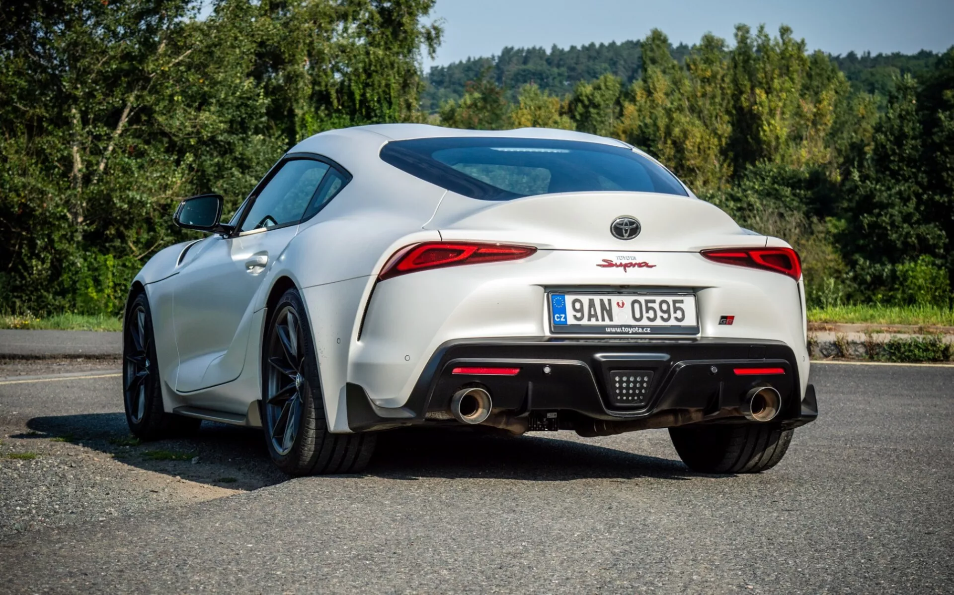 Biały Toyota Supra (GR Supra) z 2019 roku, przedstawiony z tyłu z perspektywy trzech czwartych, ze szczególnym uwzględnieniem aerodynamicznego designu, w tym tylnego spoilera, LED-owych świateł tylnych i sportowego dyfuzora, na asfaltowej powierzchni