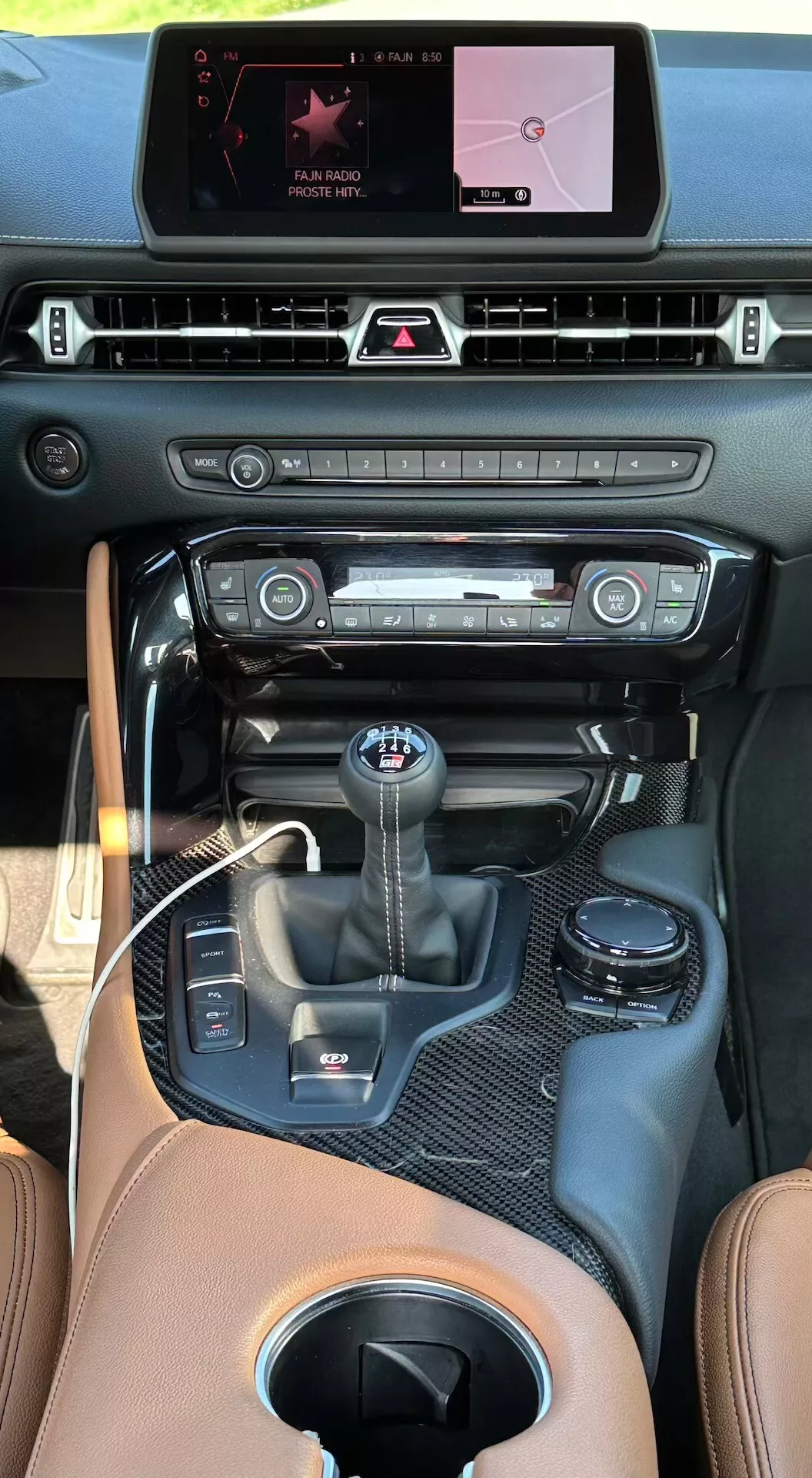 Widok wnętrza Toyota Supra (GR Supra) 2019 ze szczególnym uwzględnieniem konsoli środkowej, cyfrowego wyświetlacza systemu infotainment z nawigacją i menu medialnym, poziomych nawiewów, dwustrefowej klimatyzacji z pokrętłami i przyciskami regulacji temperatury, sportowego wybieraka biegów z funkcją manualnej zmiany, otoczonego wykończeniem przypominającym włókno węglowe, elektronicznego hamulca postojowego, obszaru ładowania USB i gniazda zasilającego, oraz wysokiej jakości tapicerki skórzanej w kolorze jasnobrązowym.