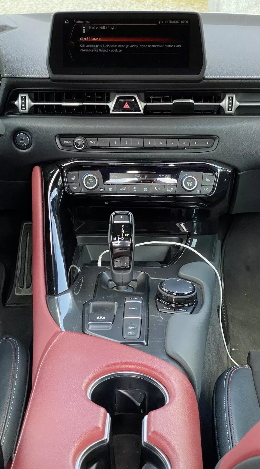 Pionowy obraz z perspektywy siedzenia kierowcy lub pasażera, ukazujący centralną konsolę i deskę rozdzielczą Toyoty Supra GR 2.0 Pure Automatic z 2019 roku. Na górze deski rozdzielczej znajduje się wyświetlacz infotainment w trybie gotowości, poniżej są dotykowe przyciski i centralny nawiew powietrza. Niżej znajdują się przyciski i pokrętła do sterowania klimatyzacją w metalicznym wykończeniu. Centralna konsola jest częściowo pokryta materiałem przypominającym czerwoną skórę z błyszczącym czarnym wykończeniem, w tym dźwignią zmiany biegów z osłoną skórzaną dla automatycznej skrzyni biegów, otoczoną przyciskami trybów jazdy, uchwytem na kubek i małym schowkiem. Podłokietnik, lekko rozmyty, również jest pokryty czerwonym materiałem. Wnętrze ogólnie sugeruje mieszankę nowoczesnej technologii i tradycyjnych fizycznych kontrolerów z wykończeniami sportowymi wysokiej klasy.