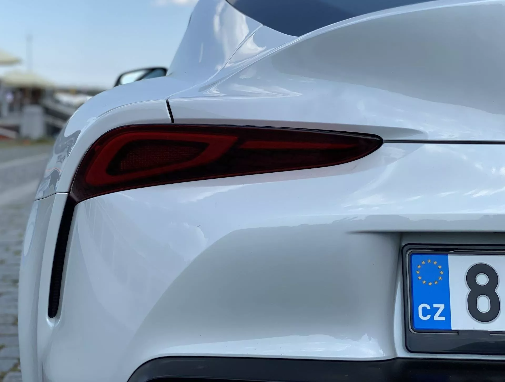 Zbliżenie częściowego widoku białego Toyota Supra 2019 (toyota-gr-supra-2-0-pure-automatic), skupiającego się na tylnej prawej sekcji z niskiego, przesuniętego kąta. Obraz ukazuje eleganckie tylne światła LED, sportowy tylny zderzak, błyszczące wykończenie lakieru i tablicę rejestracyjną zarejestrowaną w UE z Czech.