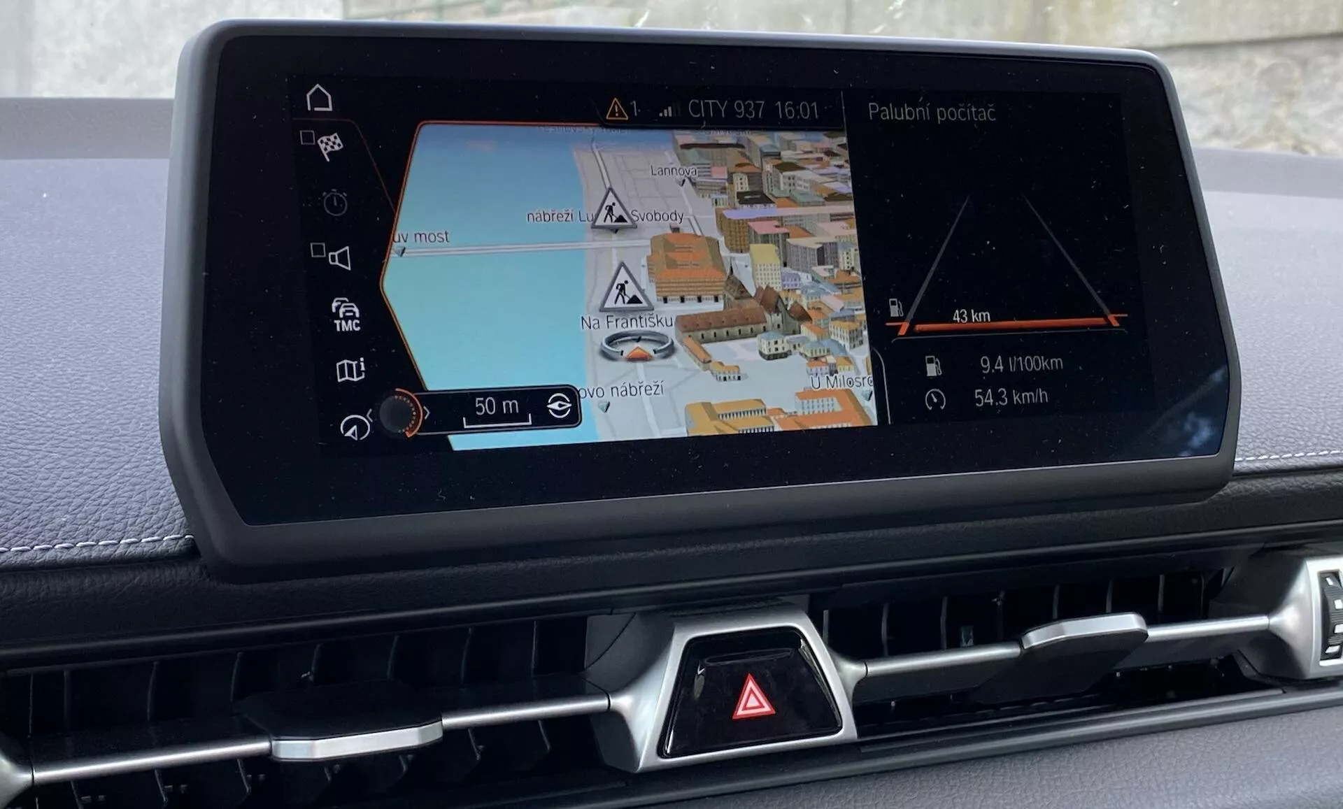 Zdjęcie ukazuje konsolę centralną Toyoty GR Supra 2.0 Pure Automatic z 2019 roku, koncentrując się na wbudowanym systemie infotainment z wyraźnym wyświetlaczem nawigacji. Zrobione z lekko zniekształconego kąta od prawej strony, ujęcie przedstawia ekran nawigacji z widokiem 3D z ulicami, budynkami i różnymi ikonami dla wskazówek nawigacyjnych i punktów zainteresowania. Linia trasy i wskaźnik pojazdu pokazują aktualne położenie samochodu, wraz ze znacznikami odległości i wskazaniami skrętów. Poniżej ekranu znajduje się rząd w większości zasłoniętych przycisków z rozpoznawalnym czerwonym trójkątem ostrzegawczym. Po bokach ekranu są kratki lub otwory, sugerujące integrację z systemem kontroli klimatu. Zbliżenie podkreśla zaawansowaną technologię samochodową i funkcje infotainment takie jak rozrywka audio, opcje łączności i ustawienia pojazdu, bez ukazywania zewnętrznej części ani reszty wnętrza.
