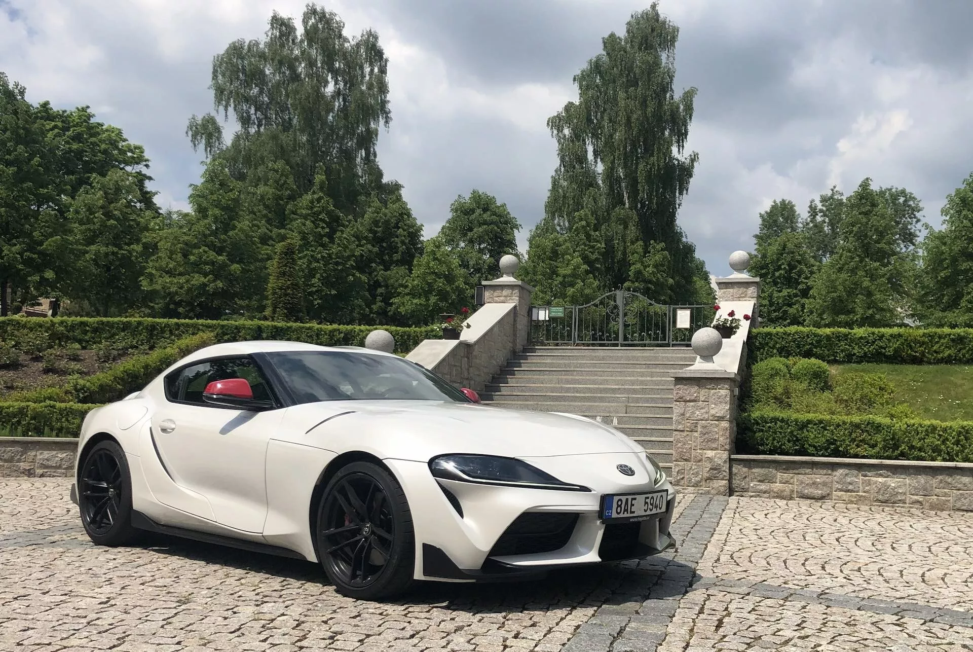 Widok trzy czwarte z przodu białego sportowego samochodu Toyota GR Supra 2.0 Pure Automatic z 2019 roku na brukowanej powierzchni, zrobiony z niskiego kąta przy jasnym niebie i roślinności w tle. Obraz podkreśla agresywną postawę samochodu, sportowe linie, ostro zaprojektowane reflektory LED, dużą przednią kratkę dla aerodynamiki i chłodzenia silnika, estetyczne i funkcjonalne wloty powietrza na masce, smukły aerodynamiczny profil boczny, zaprojektowane lusterka boczne, duże obręcze aluminiowe z hamulcami wyczynowymi i niskoprofilowe opony. Długi kaptur i kabinie umieszczonej w tylnej części podkreślają układ z przednim silnikiem i napędem na tylne koła.