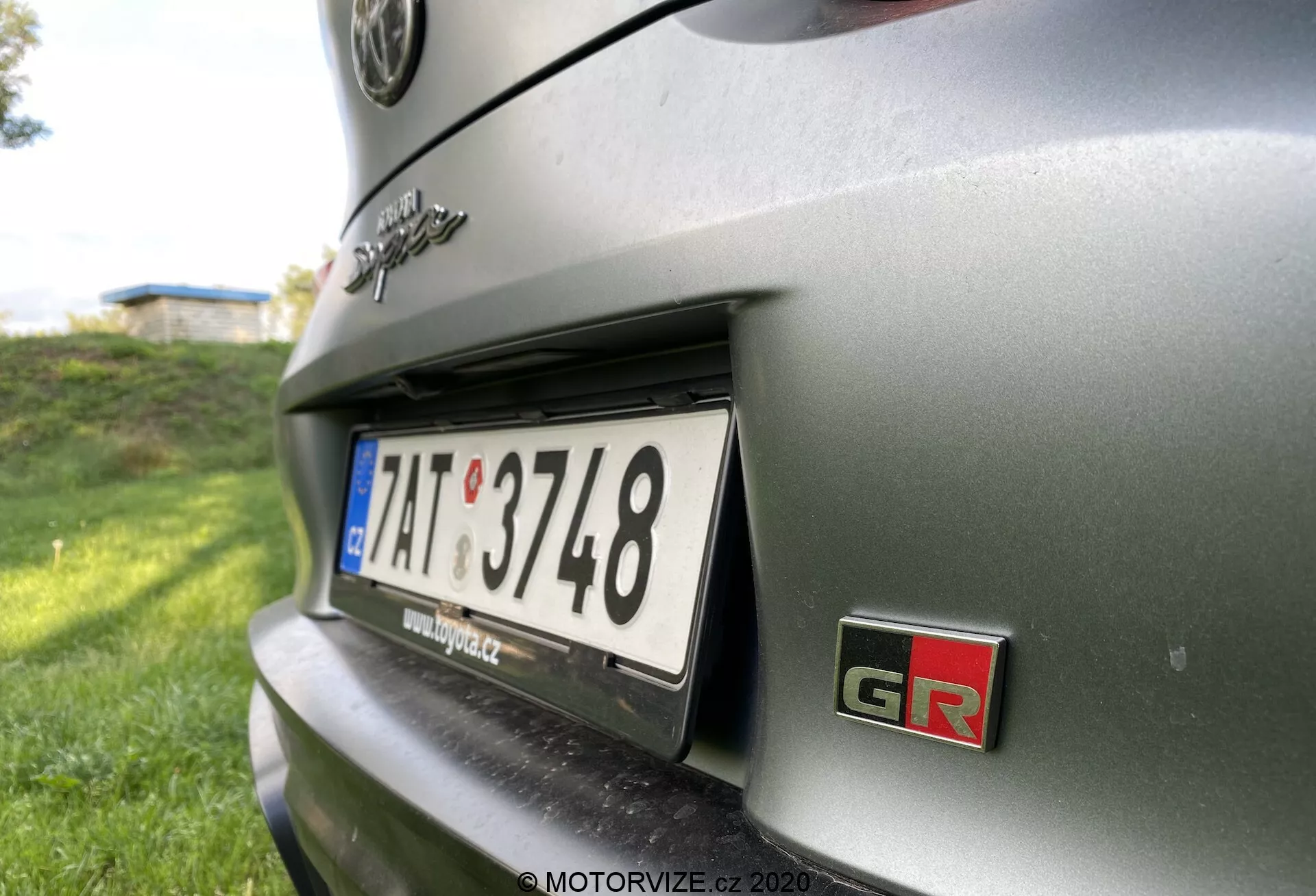 Zbliżony widok pod kątem tylnej części metalik szarej Toyoty GR Supra 2.0 Pure Automatic z roku 2019. Skupia się na obszarze tablicy rejestracyjnej i otaczających szczegółach, z naciskiem na prawą stronę tylnej części samochodu. Perspektywa jest z niskiego kąta, jakby wykonana z pozycji kucającej, patrząc lekko w górę w kierunku tyłu pojazdu. Odznaka 'GR', oznaczająca wydajnościową wersję Toyoty z linii Gazoo Racing, oraz tablica rejestracyjna europejskiego stylu są kluczowymi cechami. Głębia ostrości jest płytka, z tablicą rejestracyjną i odznaką GR w ostrym focusie, podczas gdy reszta karoserii samochodu i tło są niewyraźne. Oświetlenie sugeruje, że obraz został zrobiony na świeżym powietrzu w słoneczny dzień, z naturalnym światłem tworzącym cienie i refleksy na powierzchni samochodu.