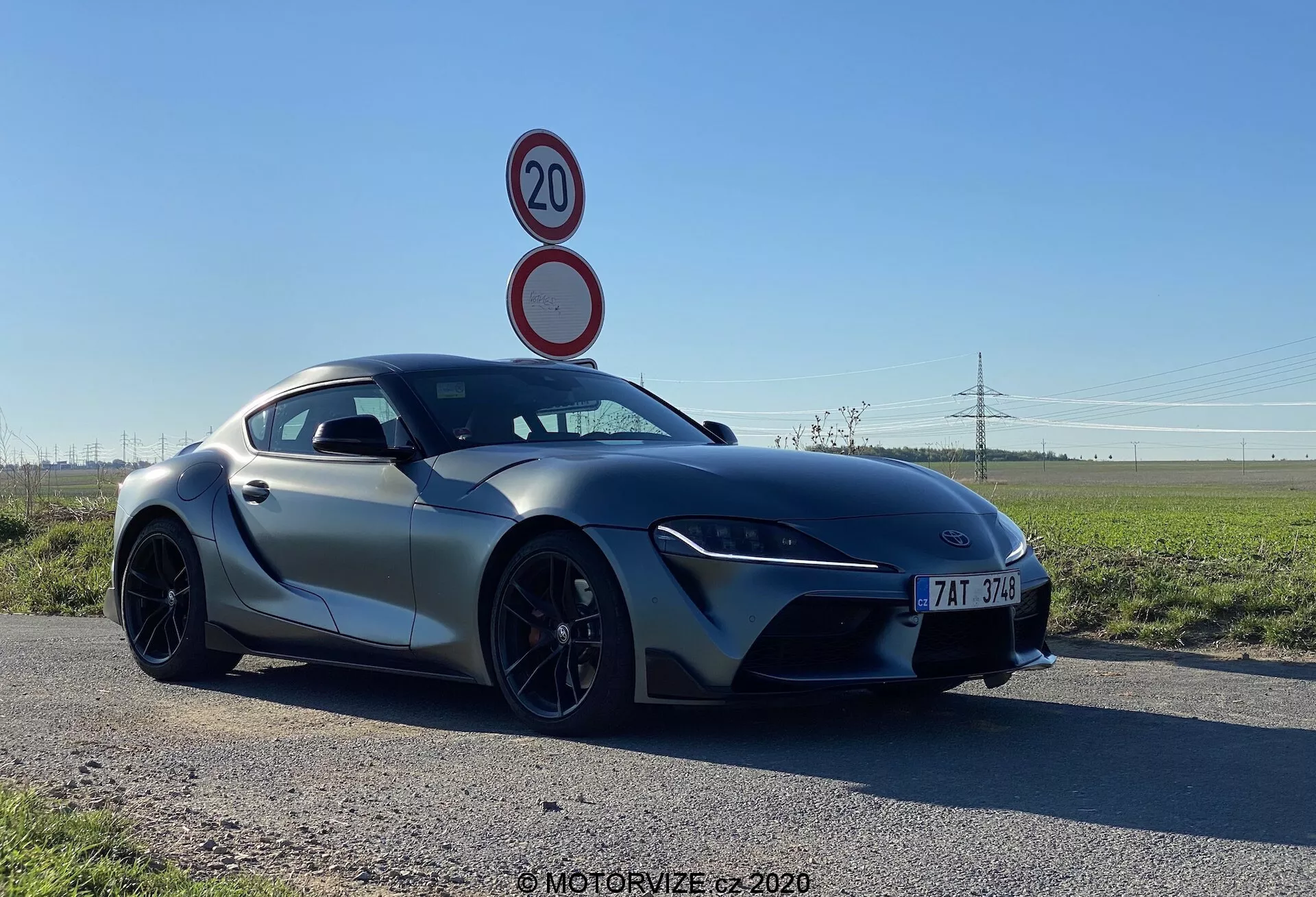 Obraz przedstawia Toyotę GR Supra 2.0 Pure Automatyczną z 2019 roku z niskiego kąta trzech czwartal z przodu, podkreślając jej smukłe, aerodynamiczne linie, wąskie reflektory LED i duże wloty powietrza z przodu. Maskę pochylono do sportowej przedniej kratki, sugerującej ułożenie silnika z przodu. Lusterka boczne są zamontowane na drzwiach, podkreślając sylwetkę typu fastback, która wzmacnia wygląd coupe. Charakterystyczne cechy obejmują wieloszprychowe czarne koła z oponami o niskim profilu i wentylowany układ hamulcowy, sugerując wysokie osiągi. Samochód znajduje się na asfaltowej drodze na tle płaskiej, wiejskiej scenerii, co podkreśla jego zaawansowaną konstrukcję.