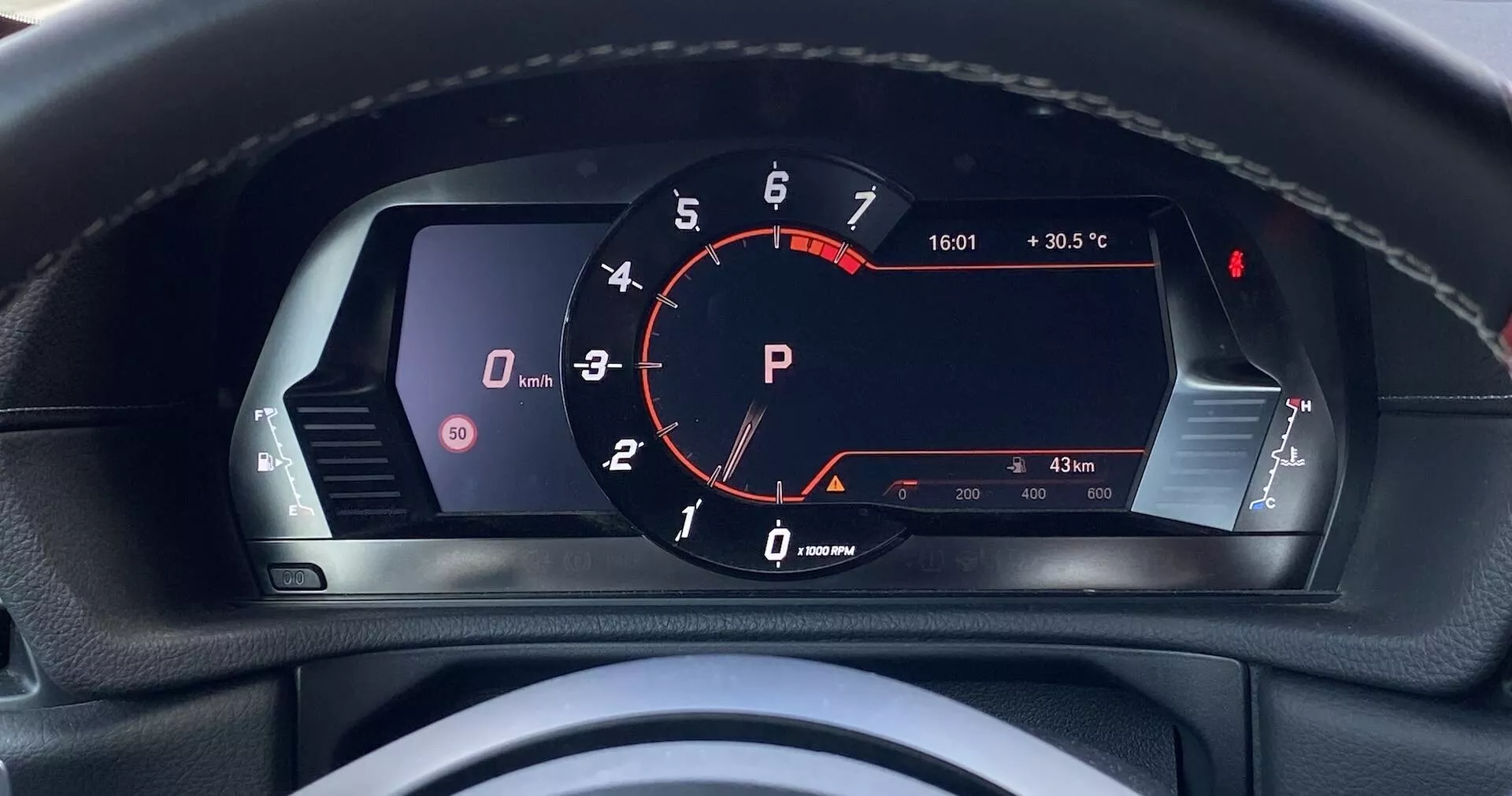 Perspektywa kierowcy na cyfrowym zestawie wskaźników Toyoty GR Supra 2.0 Pure Automatic 2019. Centralny obrotomierz z czerwoną wskazówką na '0' RPM wskazuje pracujący na biegu jałowym silnik; czerwony wskaźnik 'P' oznacza tryb parkowania. Na lewym ekranie ikona pojazdu i potencjalne kontrolki ostrzegawcze otwartych drzwi. Prędkościomierz nie jest wyraźnie widoczny, prawdopodobnie na wyświetlaczu cyfrowym. Prawa strona obejmuje wskaźniki poziomu paliwa i temperatury silnika, paliwa powyżej jednej czwartej i temperatura w normie. Dodatkowe szczegóły: temperatura na zewnątrz (30,5°C), czas (16:01) i odczyt licznika (833 km), co sugeruje niski przebieg. Widoczna górna część kierownicy sugeruje luksusowe wykończenie z detalami szwów. Perspektywa pierwszoosobowa podkreślająca zaawansowaną, nowoczesną technologię wyświetlaczy dla kierowcy.
