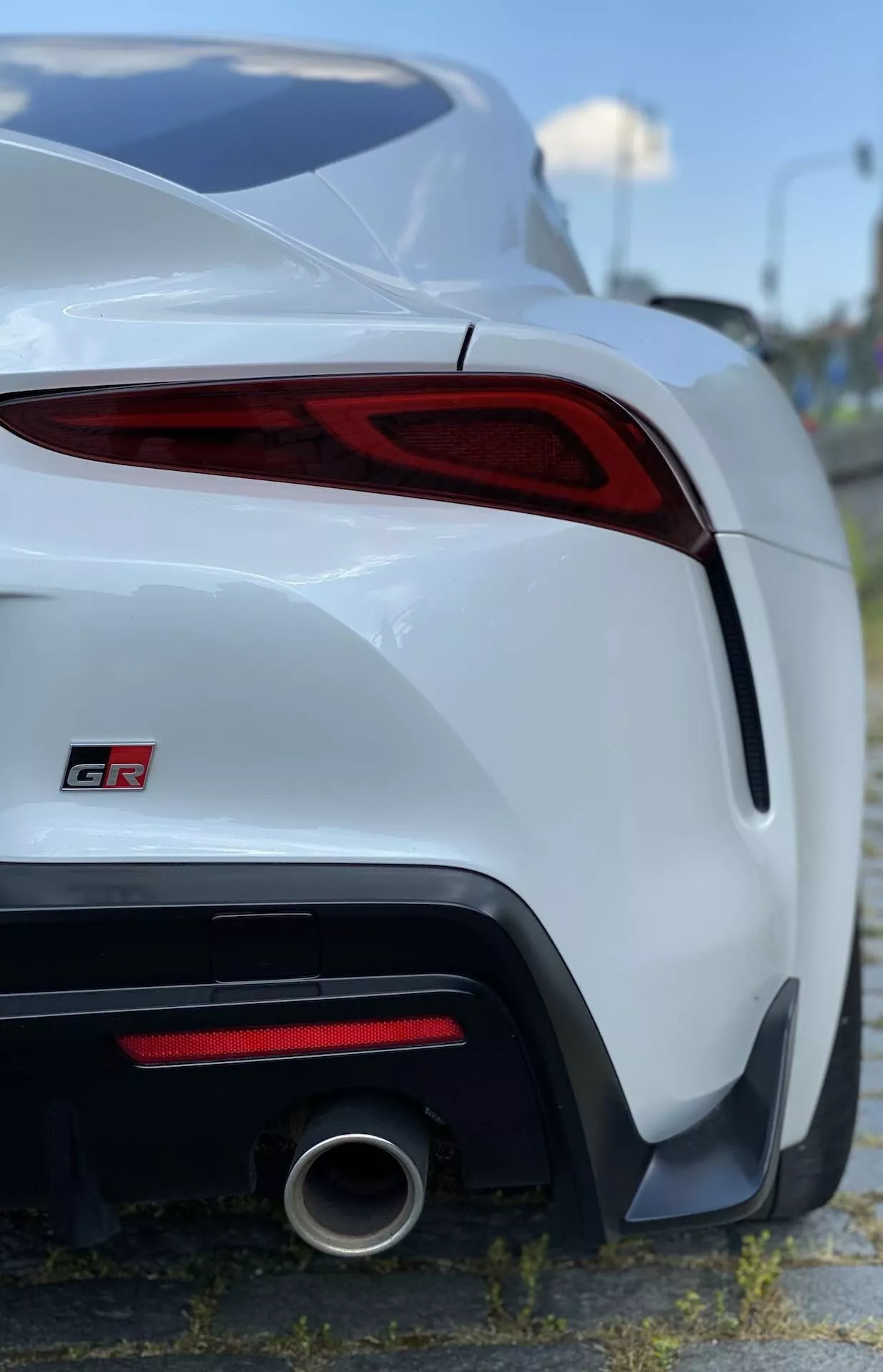 Zbliżenie tylnej części białej Toyota Supra 2019 (Toyota GR Supra 2.0 Pure Automatic) uchwycone pod niskim kątem. Lewa tylna strona ukazuje tylną część, podkreślając tylne lewe światło z ostrym designem LED, część tylnego zderzaka, pojedynczą okrągłą rurę wydechową z polerowaną metalową końcówką i trochę bocznego profilu. Wąska głębia ostrości utrzymuje samochód w centrum uwagi na lekko rozmytym tle z roślinnością i ogrodzeniem. Tylne światło zakręca wokół tylnego rogu z widocznym emblematem 'GR' na bagażniku, wskazując markę Gazoo Racing.