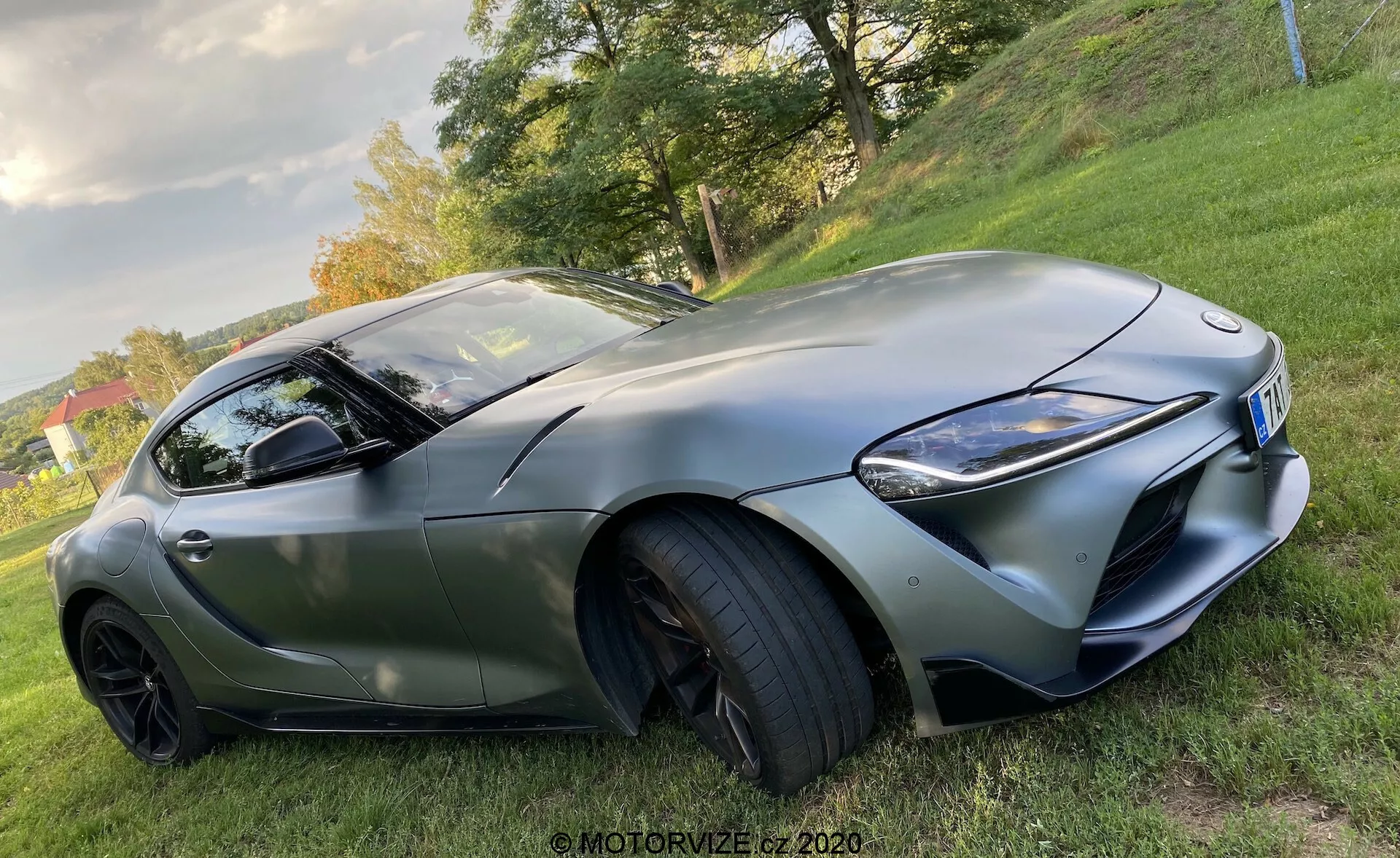 Widok z niskiego kąta na Toyota GR Supra 2.0 Pure Automatic z 2019 roku, ukazujący sportową sylwetkę z przedniego ćwiartki, podkreślającą stronę kierowcy. Obraz przedstawia aerodynamiczne kontury samochodu, wyraźny przedni grill, ostre reflektory LED, duże wloty powietrza w przednim zderzaku, rzeźbione linie maski, niskoprofilowe opony wysokiej wydajności na wieloramiennych, czarnych felgach aluminiowych, zintegrowane kierunkowskazy w lusterku bocznym i przyciemniane szyby z elegancką linią dachu i matowym wykończeniem.