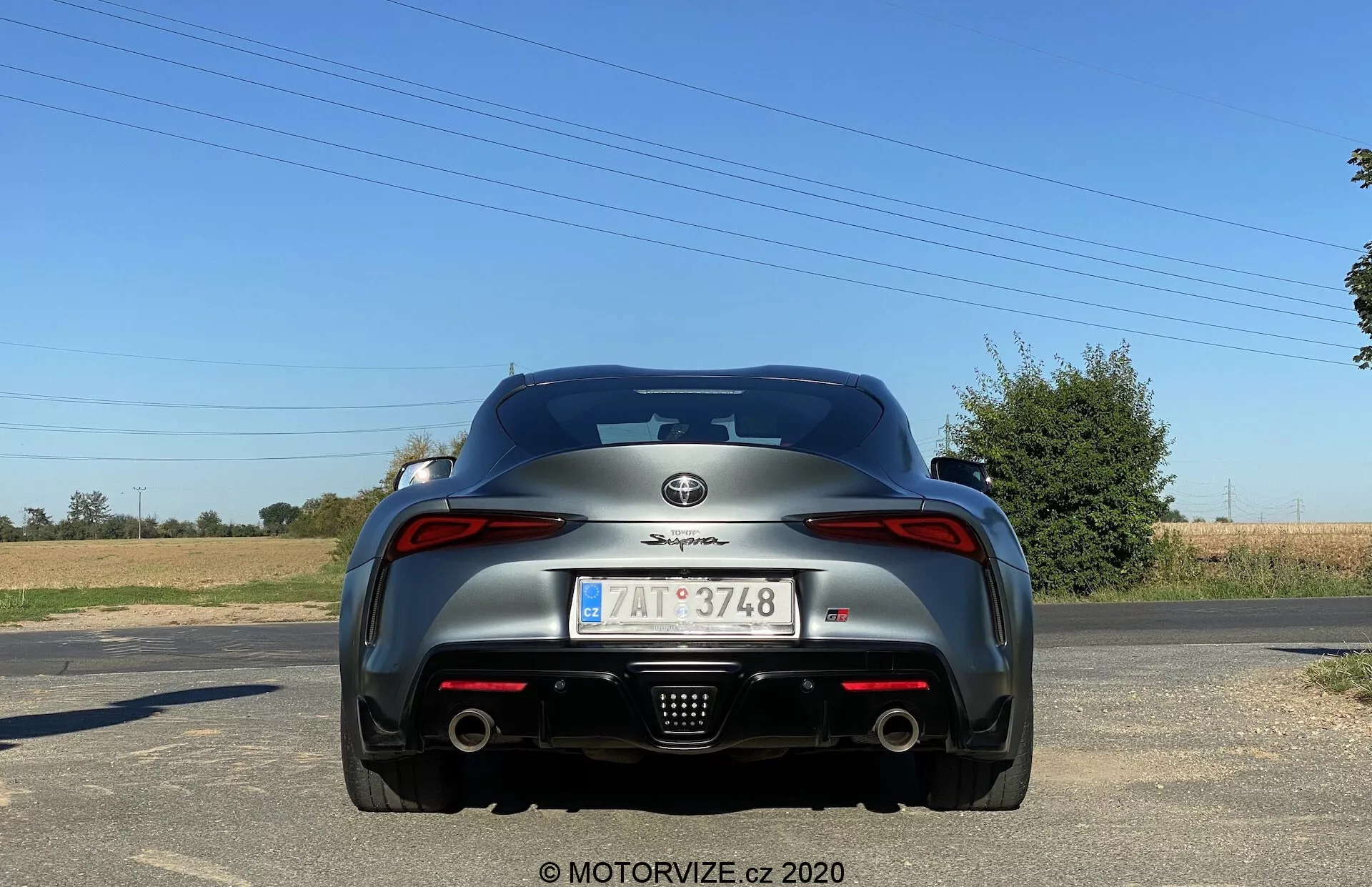 Widok od tyłu na Toyotę Suprę 2019 (Toyota GR Supra 2.0 Pure Automatic) w jasny dzień z wiejskim tłem. Zdjęcie zrobione z poziomu ziemi ukazuje tylną część nieruchomego samochodu sportowego. Widoczne elementy projektowe obejmują emblemat producenta na pokrywie bagażnika, nazwę modelu nad tablicą rejestracyjną, charakterystyczne tylne światła, podwójne końcówki wydechu, aerodynamiczne linie z zintegrowanym tylnym spojlerem oraz centralnie umieszczoną tablicę rejestracyjną z symbolem UE. Tylna szyba jest lekko zakrzywiona, a samochód jest wykończony w ciemnym, lśniącym kolorze.