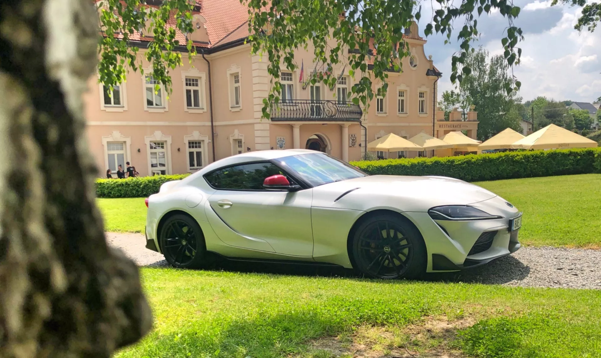 Obraz przedstawiający Toyota Suprę 2019 (Toyota-GR-Supra-2-0-Pure-Automatic) uchwycony pod niskim kątem bocznym, koncentrując się na stronie kierowcy. Samochód, pomalowany na metaliczny biały z kontrastowymi nakładkami lusterek bocznych, znajduje się około 15-20 stóp od aparatu na żwirowej ścieżce, z zadbanym trawnikiem i dużym budynkiem w tle. Widoczny jest lewy przedni kwartał pojazdu, ukazując ostre i kanciaste reflektory, wieloramienne felgi w ciemnym kolorze i duże zaciski hamulcowe, co sugeruje zaawansowaną technologię motoryzacyjną i wysokowydajny system hamulcowy.