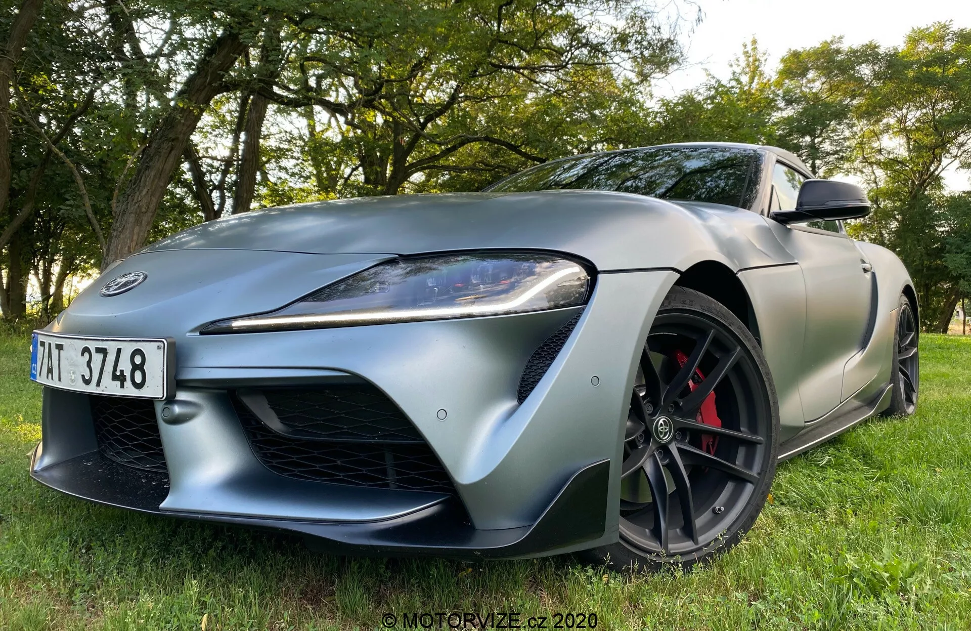 Samochód sportowy Toyota Supra 2019 widziany z niskiego, przedniego trzy czwartego kąta, zaparkowany na trawie z drzewami i liśćmi w tle. Dynamiczna i agresywna postawa podkreśla linie projektowe i aerodynamikę. Funkcje obejmują wyrazistą przednią kratkę, eleganckie kątowe reflektory LED ze światłami do jazdy dziennej, długą maskę z wyraźnymi konturami, czarne wieloszprychowe koła z kontrastującymi czerwonymi zaciskami hamulców, sportowe opony o niskim profilu, wlot powietrza na przednim błotniku i eleganckie lusterka boczne.