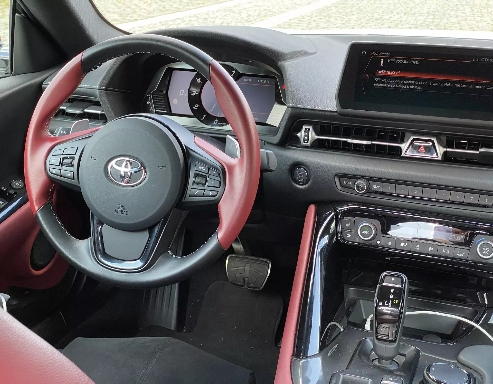 Wnętrze widziane z miejsca kierowcy w Toyocie GR Supra 2.0 Pure Automatic z 2019 r., ukazujące nowoczesną, wielofunkcyjną kierownicę z czerwonymi akcentami i różnymi przyciskami sterującymi, cyfrowy zestaw wskaźników widoczny za kierownicą oraz centralny panel z dużym wyświetlaczem systemu infotainment. Poniżej znajdują się sterowniki HVAC, a konsola centralna zawiera automatyczną dźwignię zmiany biegów i inne przyciski sterujące pojazdem. Wnętrze ma dwukolorowy schemat z materiałami skórzanymi lub podobnymi i skoordynowanymi czerwonymi akcentami.