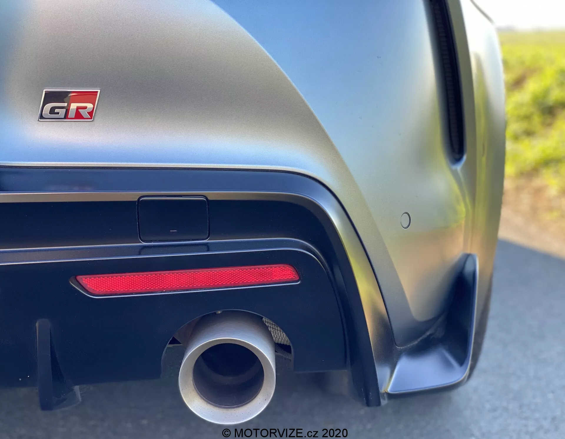 Zbliżenie bocznej perspektywy tylnego panelu samochodu Toyota GR Supra 2.0 Pure Automatic 2019, ukazujące dolne sekcje, gdzie zderzak łączy się z nadwoziem. Szczegółowy widok podkreśla nadkole, dużą okrągłą rurę wydechową, poziomy czerwony reflektor, czarną aerodynamiczną osłonę i metaliczne wykończenie, które zmienia się z ciemnych na jasne odcienie wzdłuż agresywnych linii nadwozia.
