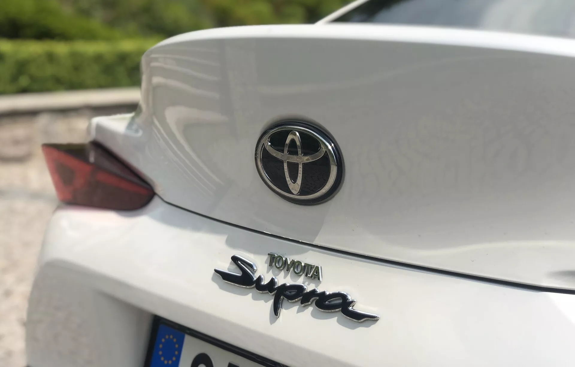 Zbliżenie tylnej części Toyota GR Supra 2.0 Pure Automatic z 2019 roku, uchwycone pod niskim do średniego kątem, koncentrujące się na emblemacie Toyoty i charakterystycznym napisie 'Supra' na białej pokrywie bagażnika. Widoczna jest także część zespołu tylnej lampy po stronie pasażera z czerwoną soczewką oraz nazwa producenta 'TOYOTA' napisane wielkimi literami. Zintegrowany spoiler dodaje sportowego wyglądu, z rozmytym tłem dzięki małej głębi ostrości.