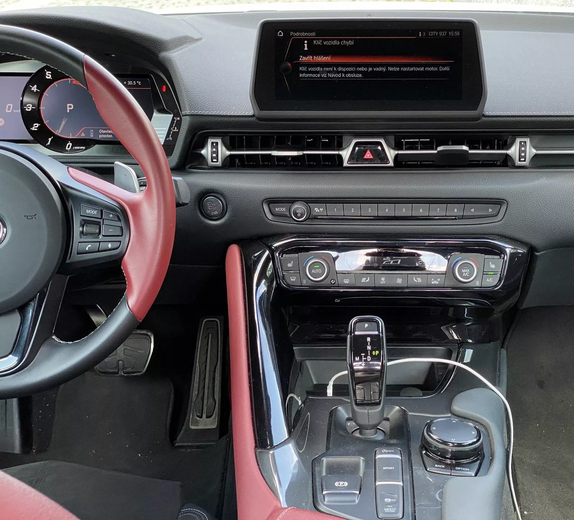 Widok z miejsca kierowcy Toyoty GR Supra 2.0 Pure Automatic z roku 2019, pokazujący konsolę środkową, wyświetlacz multimedialny, poziome kratki wentylacyjne, panel świateł awaryjnych, system kontroli klimatu z okrągłymi pokrętłami, dźwignię zmiany biegów z czarnym błyszczącym wykończeniem, przyciski do trybów jazdy i czerwoną skórzaną tapicerkę na drzwiach kierowcy.