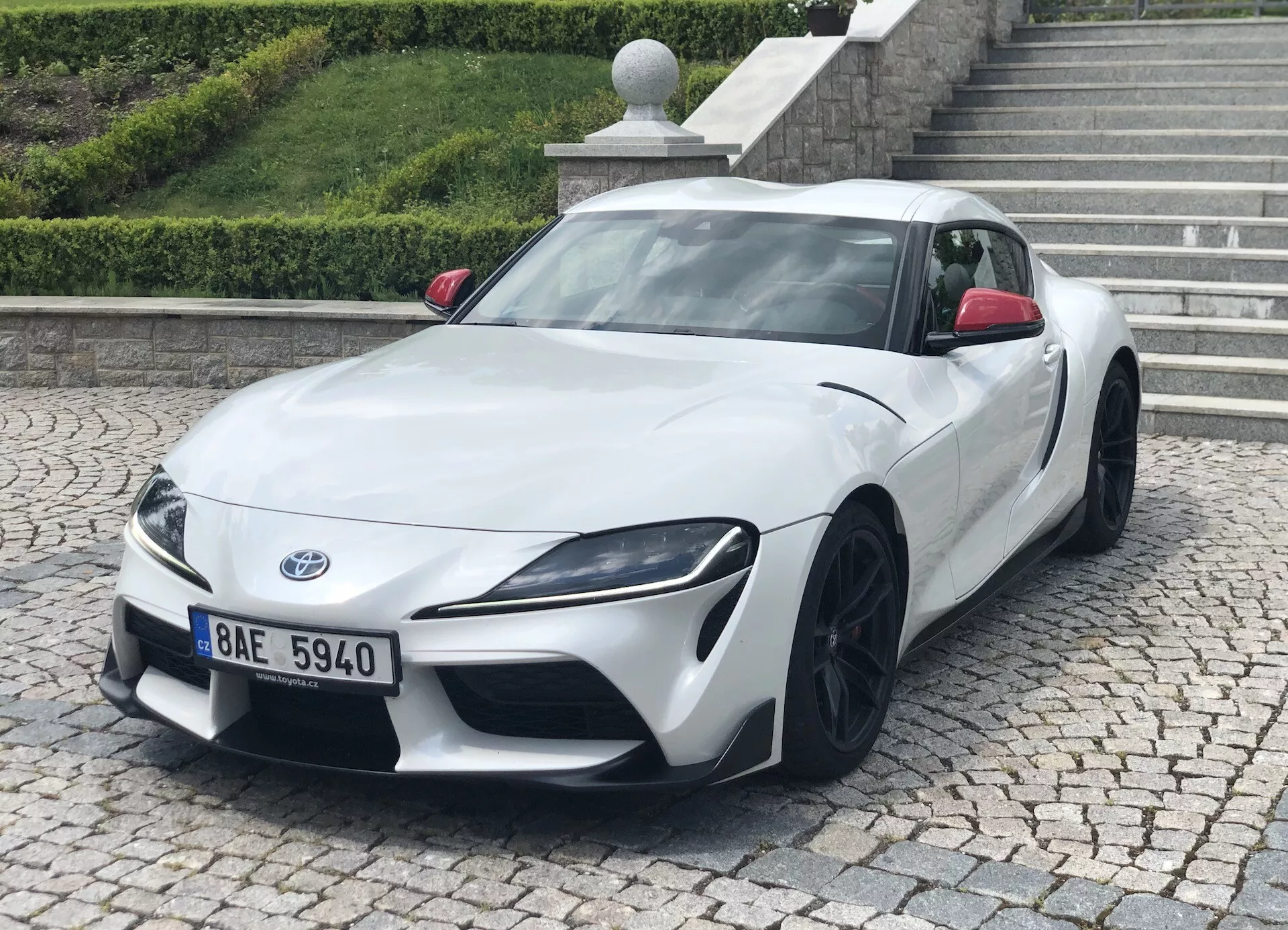 Obraz przedstawia białą Toyotę Suprę 2019 (toyota-gr-supra-2-0-pure-automatic) zrobioną z trzech czwartych przodu. Wyraźnie widać niski i szeroki wygląd auta, ostre kątowe reflektory i czarne aluminiowe felgi wieloramienne. Zdjęcie podkreśla elegancki design, aerodynamiczne kontury i sportową estetykę. Samochód sportowy jest zaparkowany na chodniku z krawężnikiem i schodami w tle, co sugeruje miejskie lub mieszkalne otoczenie.