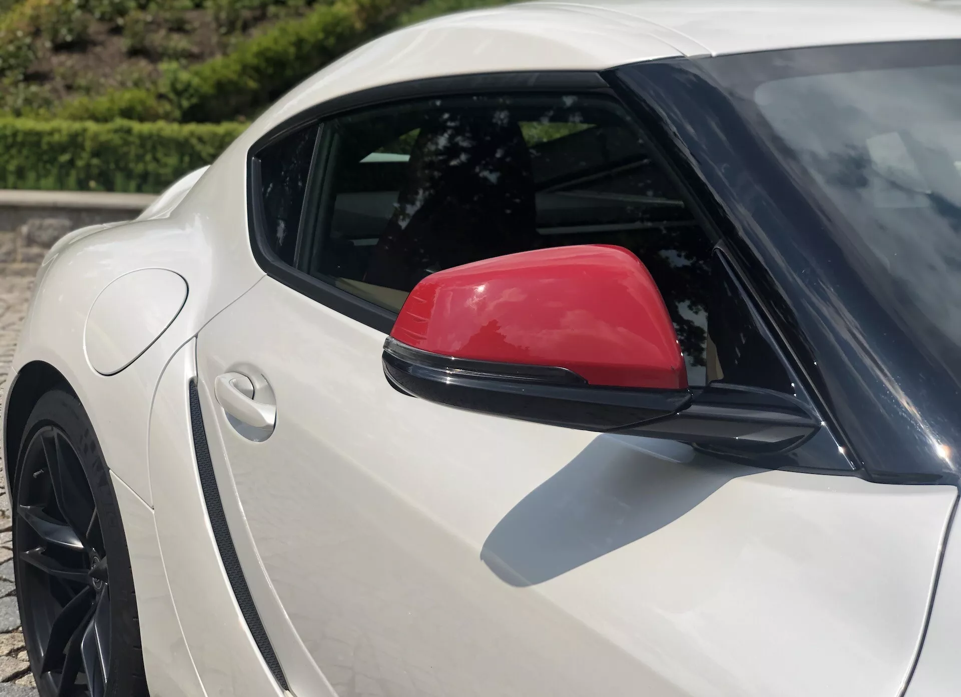 Zbliżony widok z boku na srebrną Toyota GR Supra 2.0 Pure Automatic z 2019 roku, podkreślający tylną ćwiartkę z charakterystycznym czerwonym lusterkiem bocznym i częściowo widocznym tylnym czarnym wieloramiennym aluminiowym kołem z niskoprofilową oponą; widok z podwyższonego kąta podkreśla krzywiznę tylnego błotnika i opadający dach, umieszczony w dobrze oświetlonym otoczeniu na wolnym powietrzu.