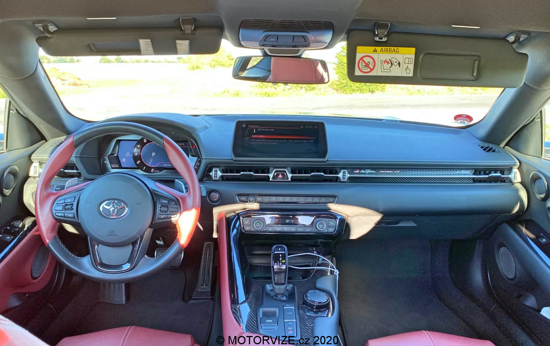 Wnętrze Toyota Supra 2019, widziane z tylnego siedzenia, ukazujące trzyramienną kierownicę z zamontowanymi kontrolerami, warstwową deskę rozdzielczą z centralnym ekranem dotykowym infotainment, smukłą kratką wentylacyjną, przyciski klimatyzacji i konsolę środkową z automatycznym selektorem biegów, elektronicznym hamulcem parkingowym i uchwytami na kubki. Czerwono-czarny wnętrze z częściowo widocznymi sportowymi siedzeniami i centralnym podłokietnikiem.