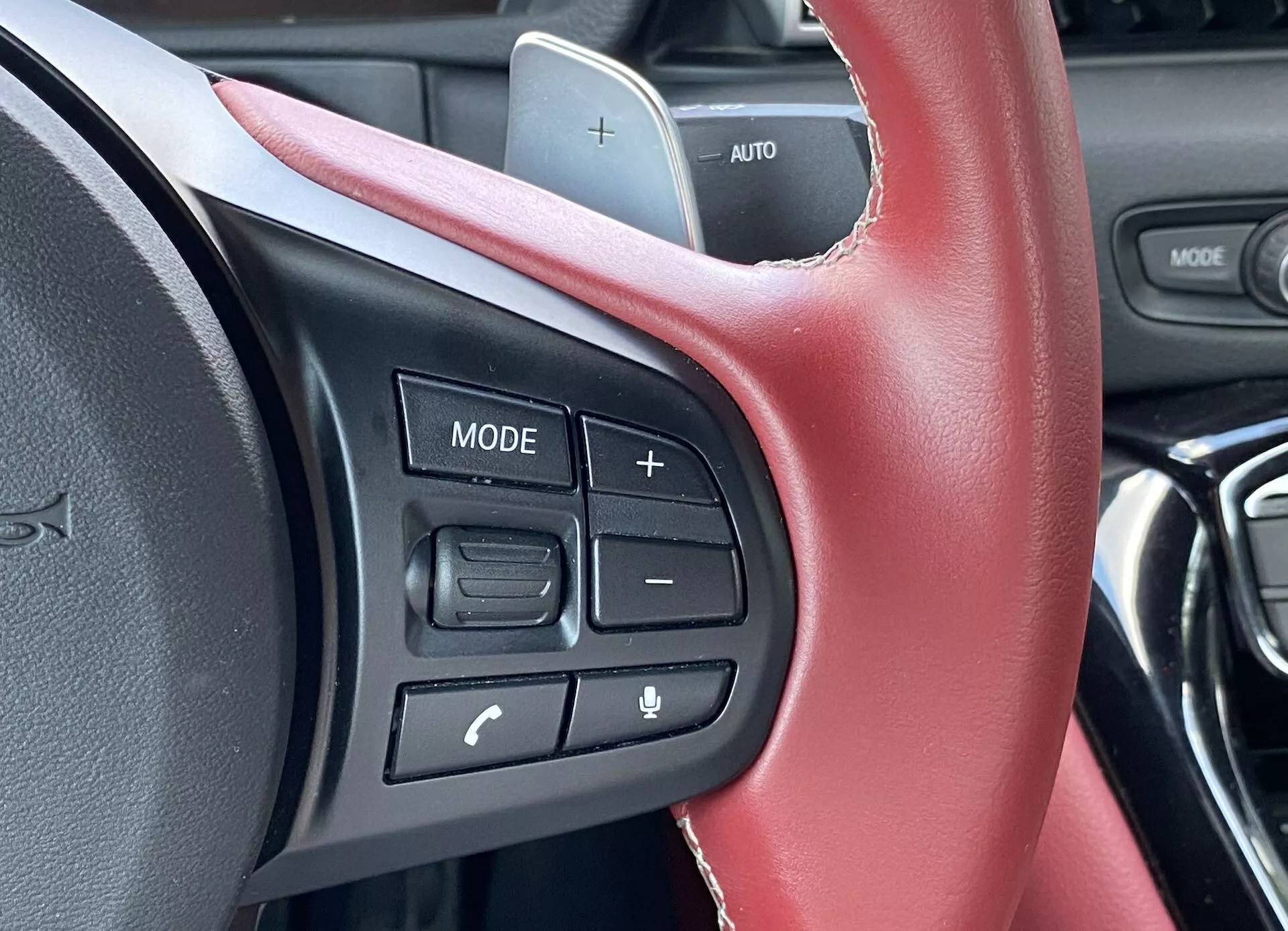 Zbliżenie sekcji kierownicy Toyoty Supry 2019 (GR Supra 2.0 Pure Automatic), skupiające się na zamontowanych na kierownicy sterownikach widzianych z perspektywy kierowcy. Skórzana kierownica z widocznymi czerwonymi szwami wskazuje na sportowe wnętrze. Dotykowe przyciski po lewej stronie obejmują przycisk 'MODE', regulatory głośności oznaczone '+' i '-', oraz dwa przyciski do obsługi telefonu i poleceń głosowych, z etykietą 'AUTO' sugerującą tempomat. Design deski rozdzielczej jest nowoczesny, ale rozmyty, bez widocznego zewnętrznego krajobrazu, co wskazuje, że samochód jest na postoju.