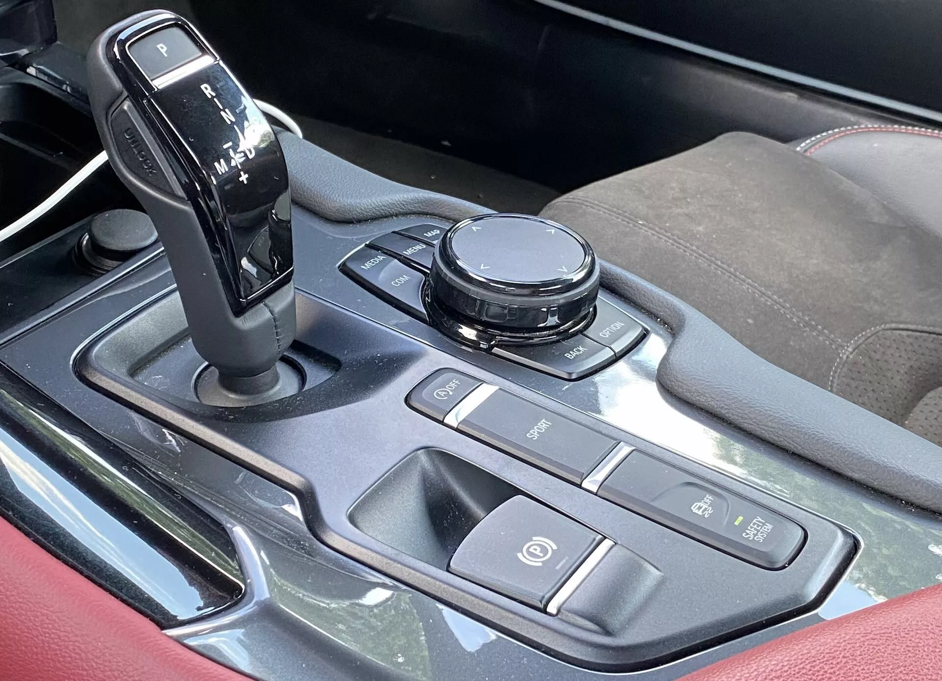 Widok z góry na centralną konsolę wnętrza 2019 Toyota Supra 2.0 Pure Automatic. Dźwignia zmiany biegów jest w pozycji parkowania, otoczona różnymi kontrolkami, w tym okrągłym pokrętłem do systemu infotainment i przyciskami do dynamiki pojazdu i skrótów infotainment. Przycisk uruchamiania/zatrzymywania silnika jest umieszczony na istotnym miejscu. Konsola posiada akcenty o metalicznym wyglądzie, polerowany metal lub wysoki połysk plastikowe i tekstylne lub skórzane podłokietniki ze szwami akcentującymi.