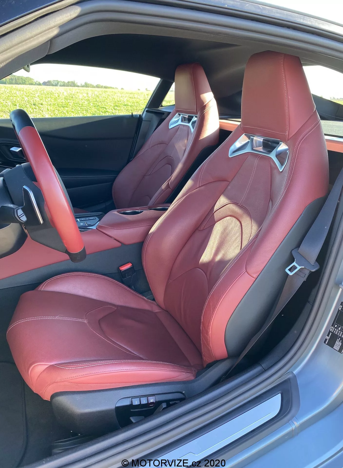 Wysokiej rozdzielczości obraz sportowego, kubełkowego przedniego siedzenia Toyota GR Supra 2019 z czerwonej skórzanej tapicerki z wyraźnymi bocznymi podparciami, zintegrowanym zagłówkiem i kontrastującym szwem. Uchwycony z zewnątrz przez otwarte drzwi, ujawnia elektroniczne sterowanie ustawieniem siedzenia i częściowy widok luksusowego wnętrza drzwi, podkreślając wygląd premium i skoncentrowany na wydajności.