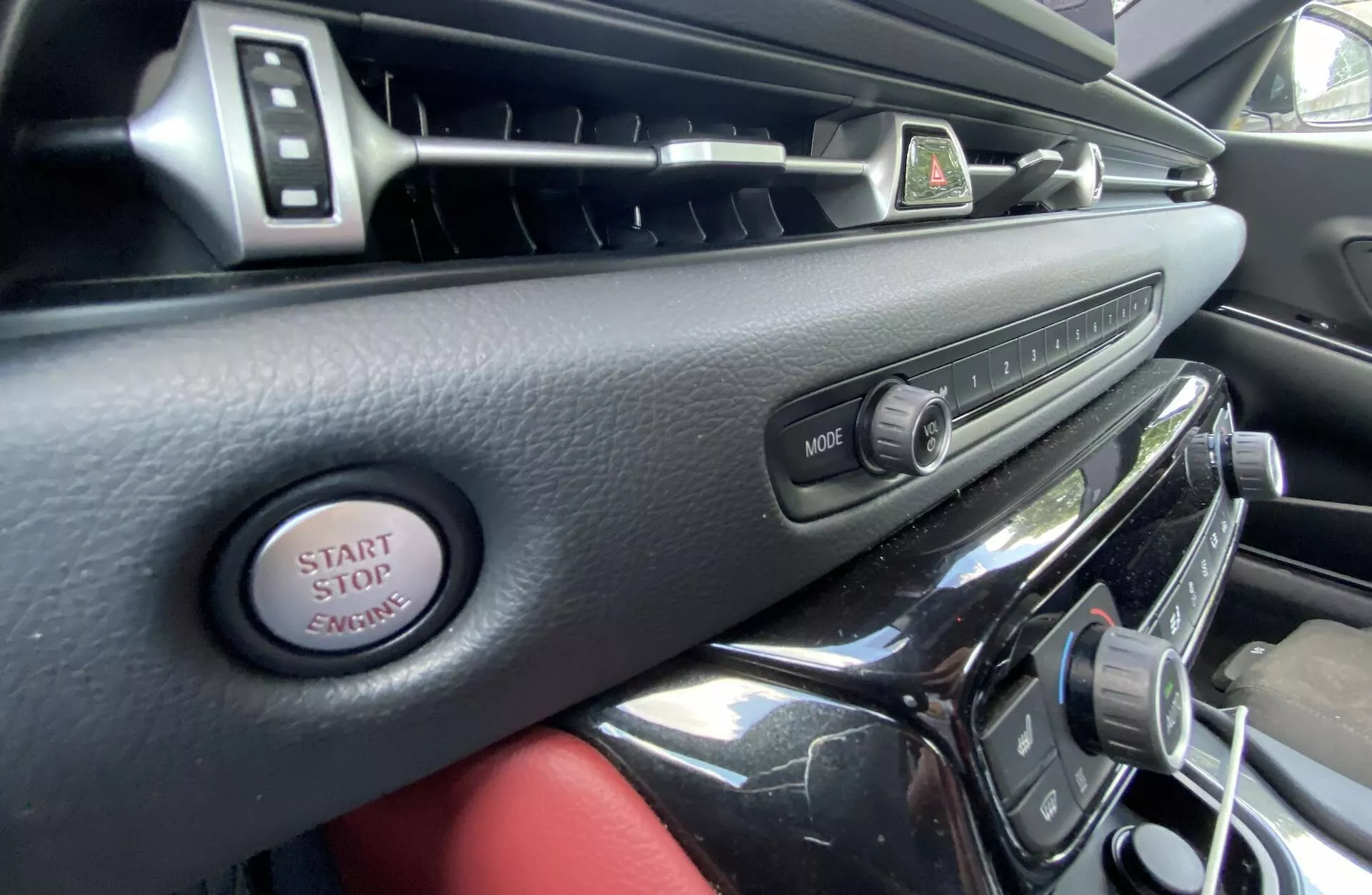 Widok wnętrza Toyota Supra 2019 z perspektywy kierowcy, prezentujący deskę rozdzielczą i konsolę środkową z chromowanymi akcentami wentylacji, przyciskiem świateł awaryjnych, przyciskiem MODE do systemu multimedialnego, przyciskiem start/stop silnika oraz połyskliwe powierzchnie, emanujące luksusem i nowoczesnością.