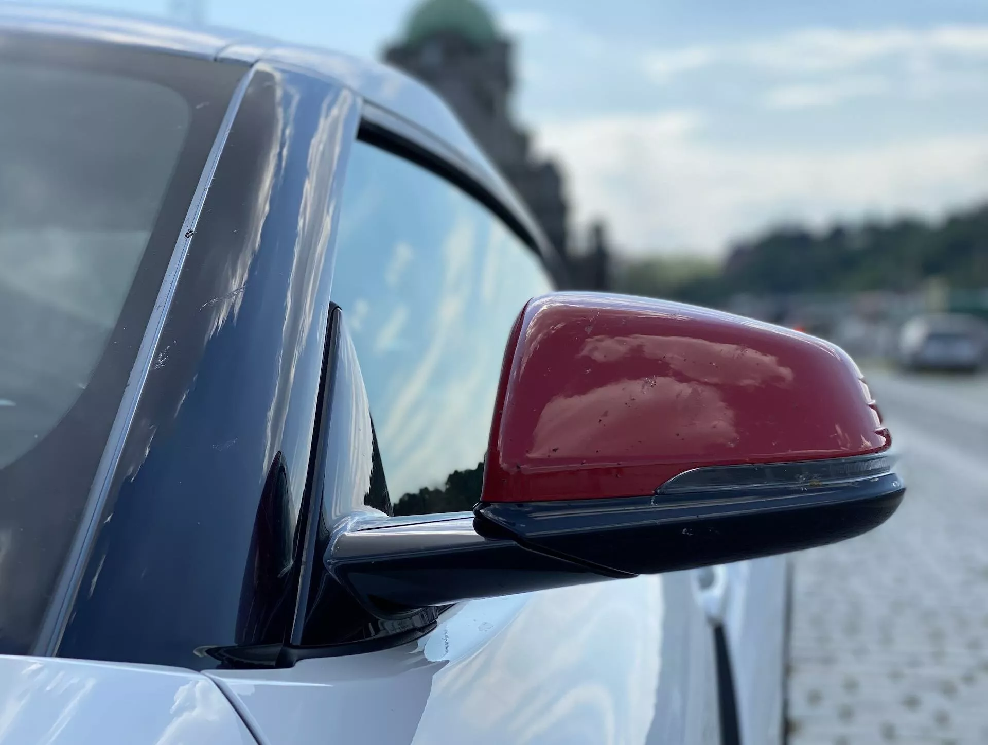 Zbliżenie bocznego lusterka Toyoty Supry 2019 z błyszczącą czerwoną pokrywą na białym nadwoziu. Aerodynamiczny design lusterka zawiera możliwe wbudowane kierunkowskazy, wskaźniki martwego pola lub funkcje auto-przyciemniania. Rozmazane tło podkreśla kontury lusterka, z elementami sugerującymi powierzchnię brukową i roślinność, co sugeruje miejskie otoczenie przydrożne.