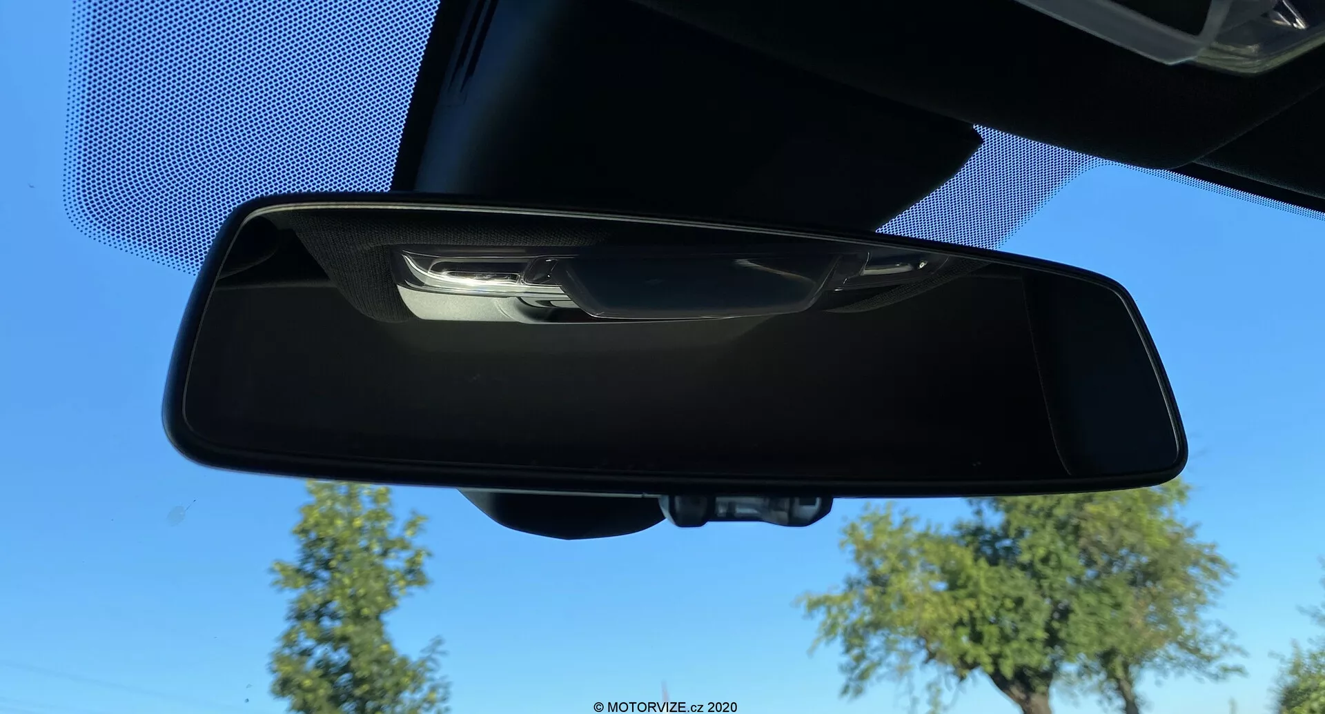 Wewnętrzne lusterko wsteczne Toyoty GR Supra 2.0 Pure Automatic z 2019 roku, widziane z dolnej przedniej perspektywy z odbiciem pokazującym niebieskie niebo, drzewa i część sufitu samochodu. Lusterko ma czarną plastikową ramę i ręczny mechanizm przechylny; nie są widoczne zaawansowane funkcje, takie jak automatyczne ściemnianie czy wbudowane wyświetlacze.