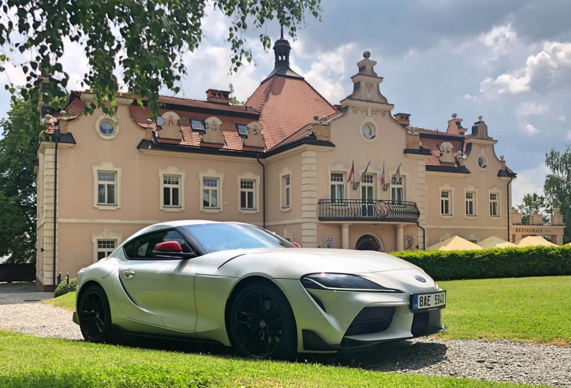 Fotografia z niskiego kąta przedstawiająca Toyotę GR Supra 2.0 Pure Automatic z 2019 roku, skupiająca się na profilu bocznym kierowcy. Smukłe, aerodynamiczne linie i rzeźbiona karoseria sugerują sportowy design. Cechy obejmują dwukolorowy schemat kolorów z głównym korpusem w kolorze srebrnym/jasnoszarym i czarnymi akcentami, charakterystyczne reflektory LED, duże wloty powietrza, czarne lusterka boczne, wpuszczoną klamkę drzwi i duże, wieloramienne czarne koła z niskoprofilowymi oponami. Czerwona osłona lusterka bocznego dodaje kontrastującego akcentu koloru. Samochód zaparkowany jest na żwirowej powierzchni przed zabytkowym budynkiem, pod częściowo zachmurzonym niebem, co uwydatnia jego krzywizny i elementy konstrukcyjne.