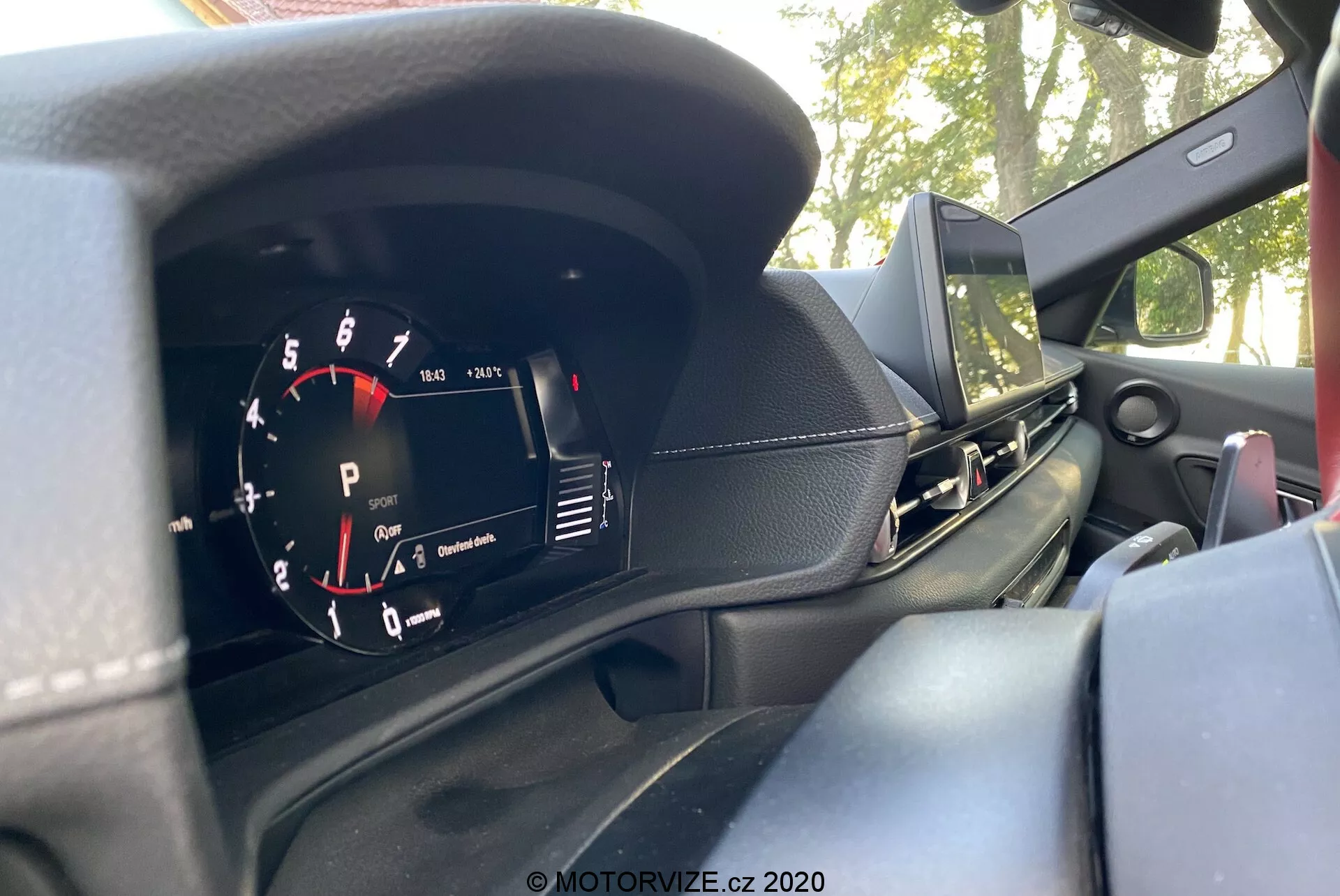 Wnętrze Toyota GR Supra 2.0 Pure Automatic z 2019 roku, skupiające się na zestawie wskaźników za kierownicą, zrobione z perspektywy siedzenia kierowcy. Analogowy obrotomierz, oznaczony w krokach co 1000 RPM z czerwonym polem zaczynającym się przed 7000 RPM, jest widoczny po lewej stronie. Po prawej stronie znajduje się wyświetlacz cyfrowy pokazujący 'P' dla trybu parkowania. Deska rozdzielcza ma czarną teksturowaną powierzchnię, z widoczną częścią ekranu infotainmentu, nawiewem powietrza po stronie pasażera oraz możliwym zamontowanym na desce rozdzielczej głośnikiem. Na obrazie częściowo widoczna jest kierownica sugerująca sportowy design. Na zewnątrz, przez przednią szybę widać drzewa i niebo, co wskazuje na to, że samochód jest zaparkowany na zewnątrz.