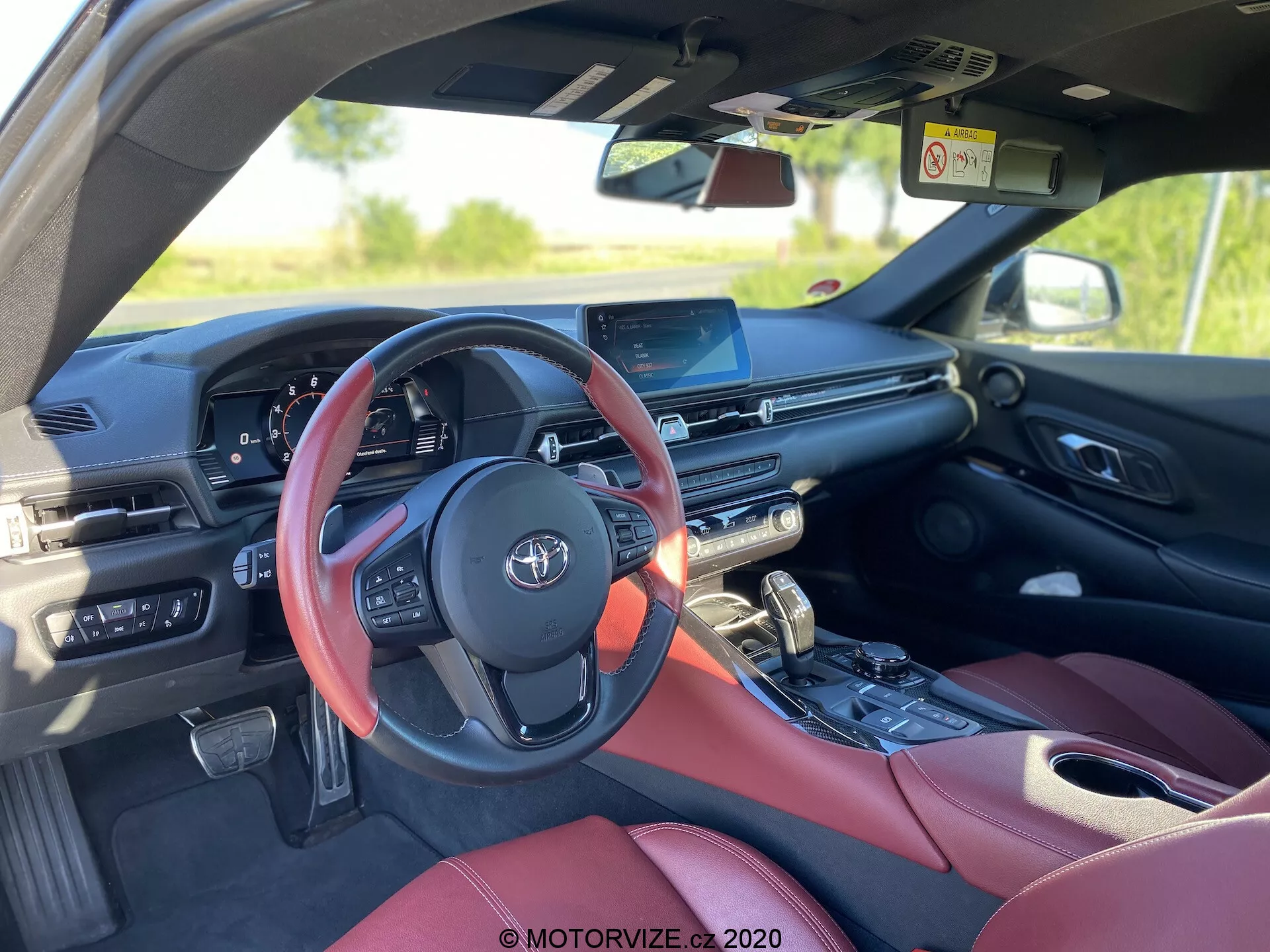Widok wnętrza Toyoty GR Supra 2.0 Pure Automatic z 2019 roku, przedstawiający lekko niewycentrowaną perspektywę tylną z tylnego siedzenia po stronie pasażera. Wyróżnia się czerwonym kierownicą z zamontowanymi sterownikami, cyfrowym panelem instrumentów, centralnym ekranem dotykowym systemu infotainment, częściowo widocznym sterowaniem klimatyzacją, dźwignią zmiany biegów z pobliskim pokrętłem sterującym oraz luksusowymi, czerwonymi skórzanymi siedzeniami, sugerującymi pakiet premium. Obraz podkreśla nowoczesną technologię samochodową i funkcje, takie jak system infotainment, cyfrowy wyświetlacz i sterowniki na kierownicy.
