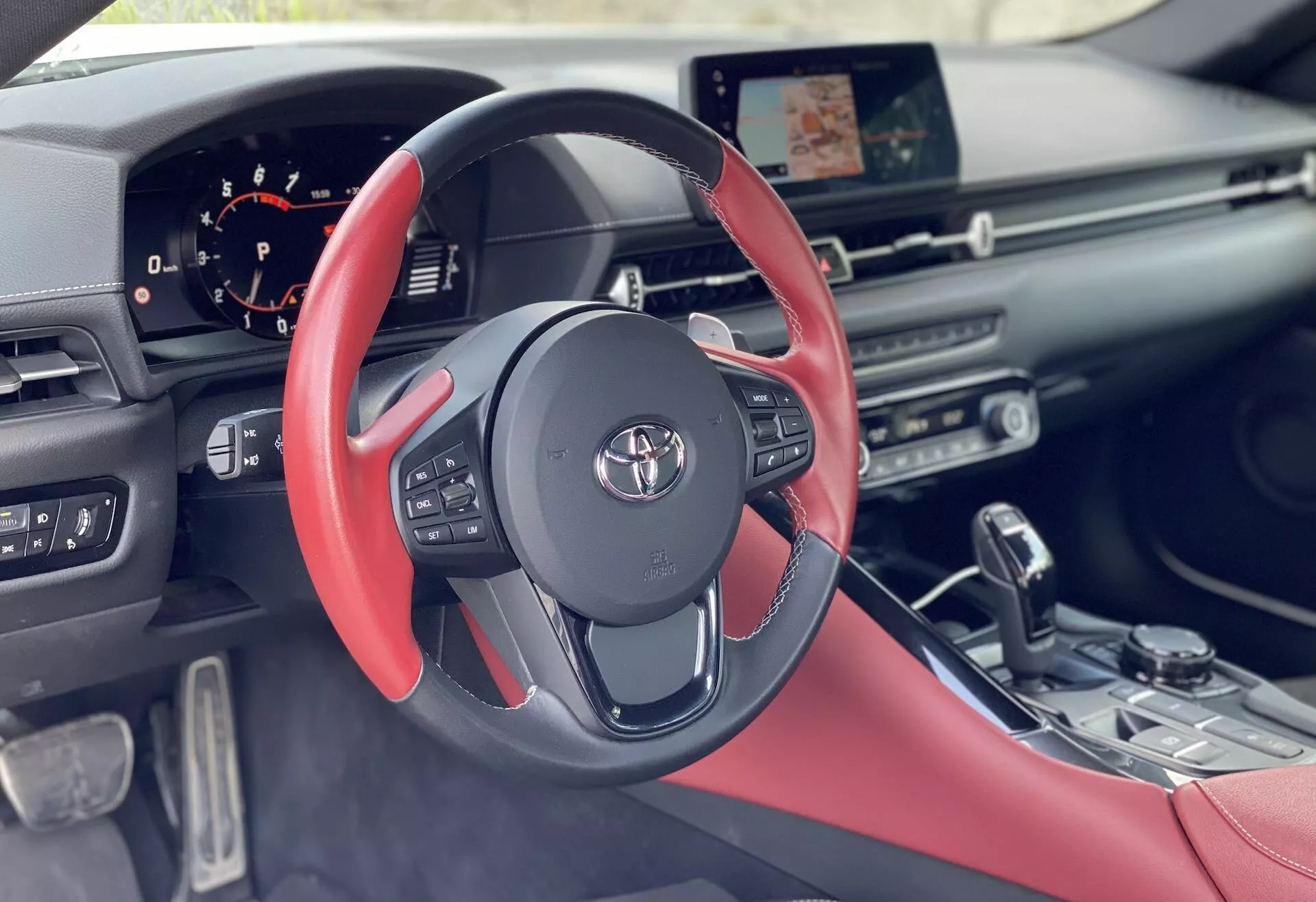 Widok wnętrza z perspektywy kierowcy Toyota GR Supra 2.0 Pure Automatic 2019. Ujęcie na poziomie oczu podkreśla sportową kierownicę z płaskim dnem i czarno-czerwonymi akcentami. Cyfrowy zestaw wskaźników z tyłu pokazuje informacje o pojeździe. Dotykowy ekran systemu multimedialnego znajduje się nad elegancką deską rozdzielczą z liniowymi otworami wentylacyjnymi. Konsola centralna zawiera metaliczną dźwignię automatyczną. Czerwone, prawdopodobnie skórzane, fotele dopełniają nowoczesny, ergonomiczny design samochodu.