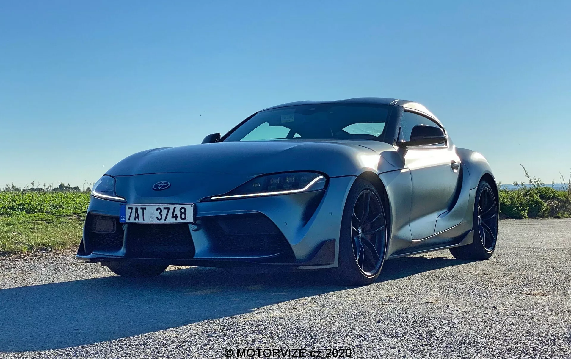 Na tym zdjęciu widać samolubnego Toyotę GR Supra 2.0 Pure Automatic z 2019 roku uchwyconego pod niskim kątem, w ciągu dnia przy bezchmurnym niebie. Zdjęcie podkreśla agresywną i sportową sylwetkę pojazdu z lewego przedniego narożnika, ukazując aerodynamiczny wygląd z widocznymi elementami takimi jak przednie reflektory, grill, przedni zderzak, drzwi kierowcy, boczne lusterko, część tylnego błotnika, kontury maski i przednie koło z aluminiową obręczą. Samochód wypełnia większość kadru, co wskazuje na bliskie podejście, które podkreśla jego nowoczesny design, ostre reflektory LED, dużą przednią kratkę i matowe wykończenie lakieru.