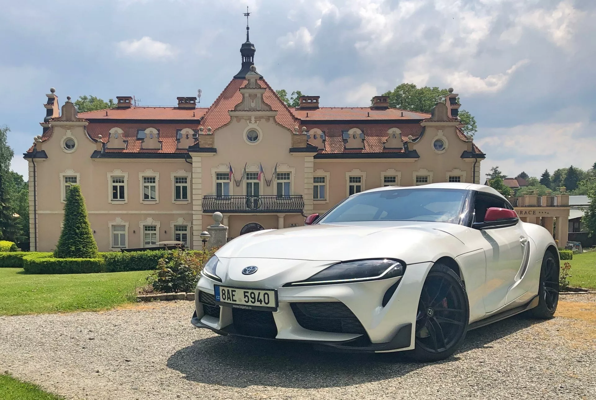 Strzał pod kątem z przodu białego samochodu sportowego Toyota GR Supra 2.0 Pure Automatic 2019 z eleganckimi, kanciastymi reflektorami LED, wyraźnym przednim zderzakiem z wbudowanymi wlotami powietrza, niską, szeroką przednią kratownicą z charakterystycznym emblematem, dużymi wieloramiennymi czarnymi felgami aluminiowymi, aerodynamicznie zaprojektowanymi lusterkami bocznymi, subtelnymi konturami maski i lekkim podwyższeniem z tyłu wskazującym na spojler. Samochód jest zaparkowany na żwirowej powierzchni z trawiastym obszarem na pierwszym planie, w słoneczny dzień, z ozdobnym budynkiem w tle, co sugeruje luksusowe lub historyczne otoczenie.