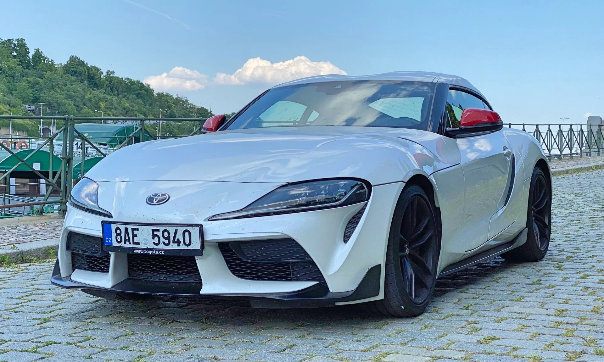 Niskokątny widok przodu białego Toyoty Supra 2.0 Pure Automatic z 2019 roku, ukazujący kluczowe elementy takie jak kątowe reflektory LED, wyraźna przednia kratka, sportowy zderzak z wbudowanymi wlotami powietrza, częściowo widoczny znak Supra, przedni splitter, charakterystyczne linie maski, czarne felgi aluminiowe wieloramienne odsłaniające wydajny układ hamulcowy oraz aerodynamiczne lusterka boczne. Linia dachowa coupe opada do kompaktowej tylnej części w świetle dziennym, w pobliżu zbiornika wodnego z metalową strukturą w tle.