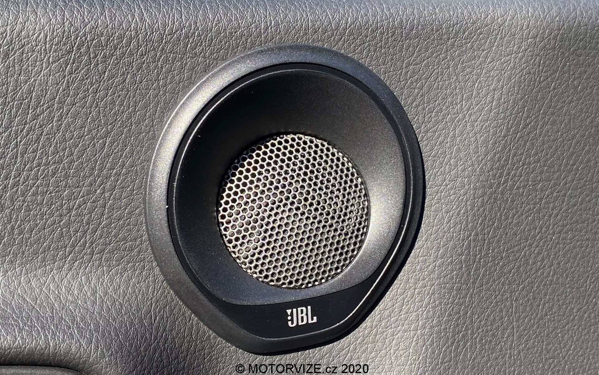 Zbliżenie głośnika samochodowego Toyota Supra 2019, wyprodukowanego przez firmę JBL, pokazujące bezpośredni widok z przodu z lekko nachylonym kątem. Okrągły głośnik ma charakterystyczną kratkę w kształcie plastra miodu, zoptymalizowaną dla czystej produkcji dźwięku. Widoczna jest okrągła ramka z kontrastowego materiału, prawdopodobnie gumy lub plastiku. Głośnik jest zamontowany na teksturowanej powierzchni przypominającej skórę, podkreślając luksusowe wrażenie. Logo JBL jest umieszczone w dolnej części, wskazując na koncentrację na wysokiej jakości systemach dźwiękowych.