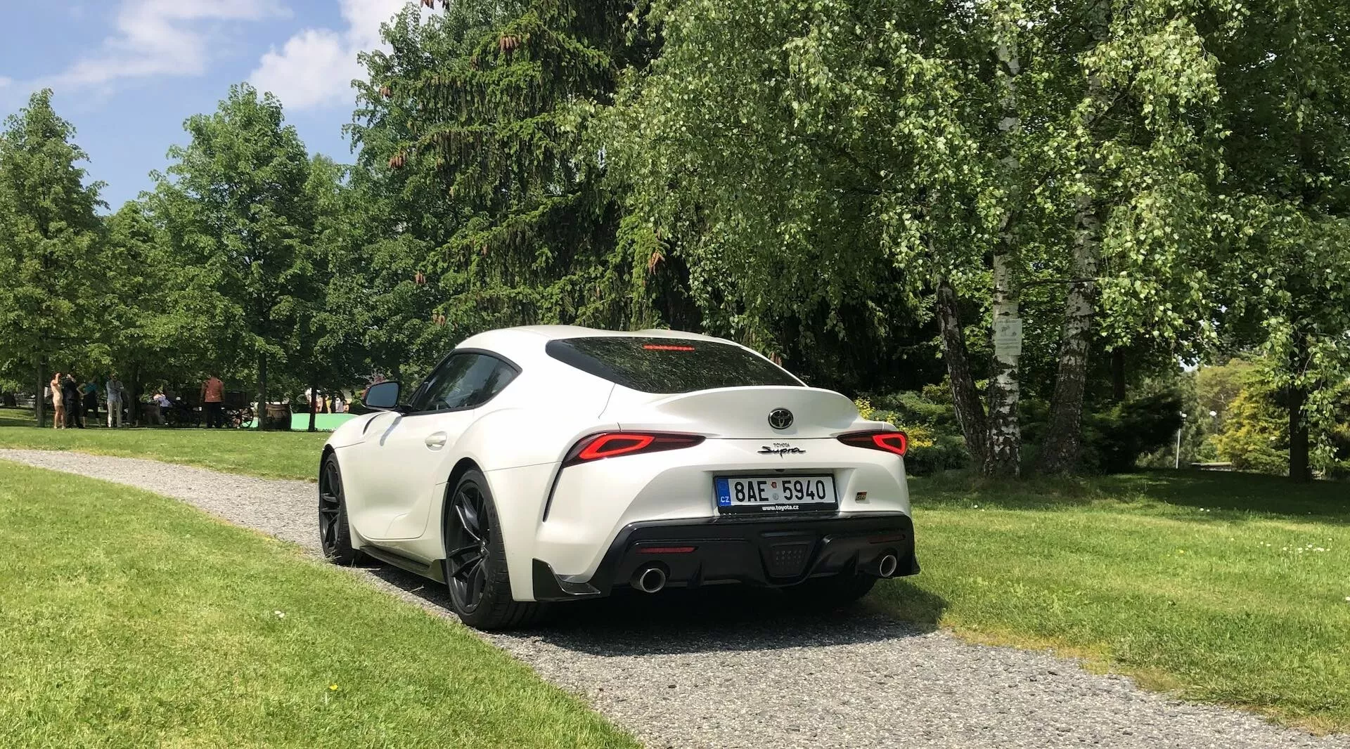 Widok od tyłu z boku na Toyota GR Supra 2.0 Pure Automatic z 2019 roku, w kolorze białym z kontrastującymi czarnymi elementami, zaparkowany na żwirowej ścieżce w otoczeniu parku. Obraz uchwytuje cechy projektowe, takie jak tylny spoiler, podwójne końcówki wydechu, wąskie tylne światła LED, sportowe felgi aluminiowe oraz lusterka boczne w kolorze nadwozia z zintegrowanymi kierunkowskazami.