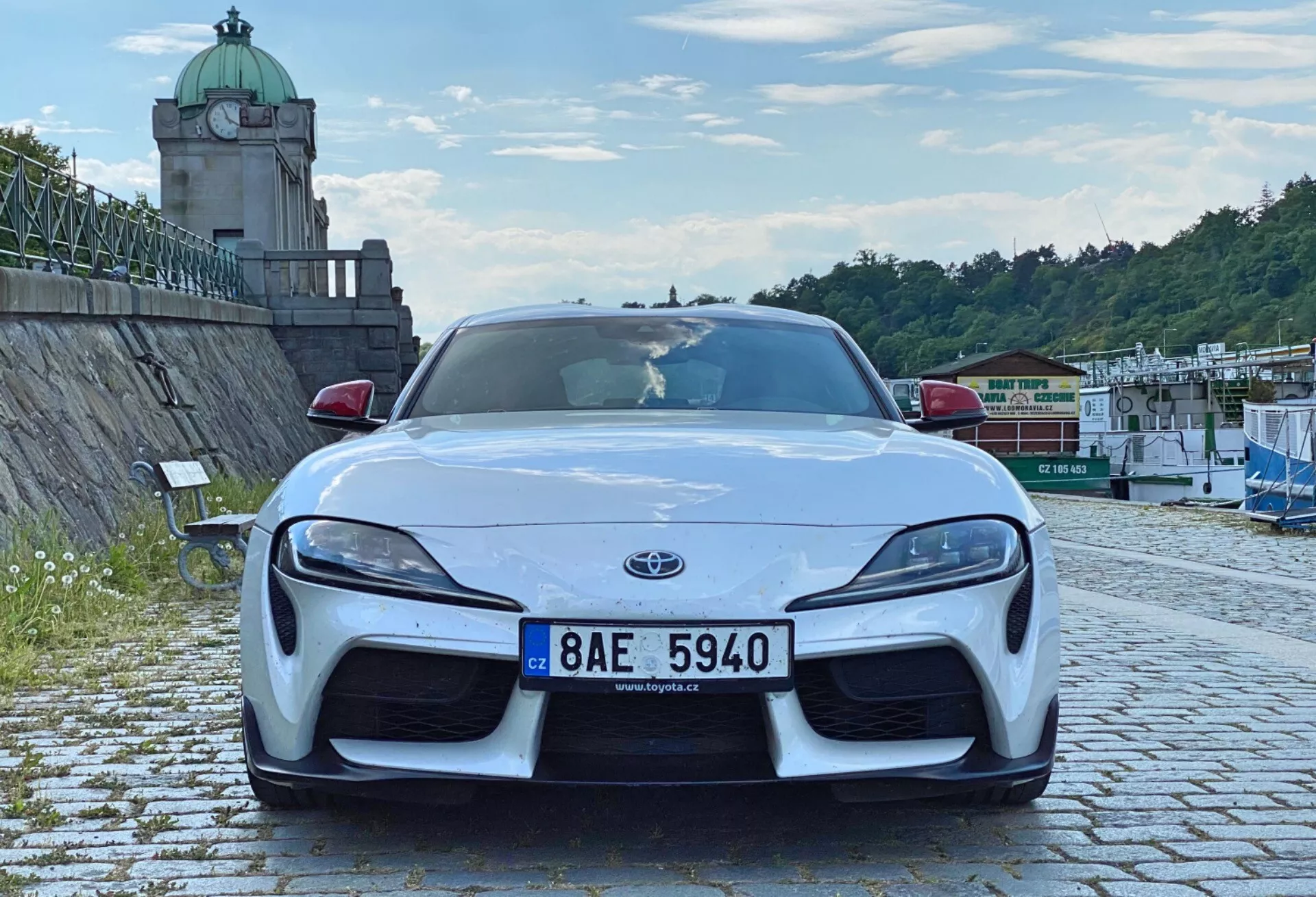 Ujęcie przodu nowoczesnego samochodu sportowego, konkretnie Toyota Supra 2019. Samochód jest wyśrodkowany w kadrze i sfotografowany z niskiego kąta, pokazując jego agresywną sylwetkę i sportowe linie. Obraz podkreśla przedni zderzak, maskę, błotniki, emblemat Toyoty, eleganckie reflektory LED, duże wloty powietrza w zderzaku oraz splitter przedni. Wyrzeźbiona maska ​​i błotniki sugerują układ napędu na tylne koła o wysokiej wydajności. Samochód zaparkowany jest na brukowanej nawierzchni w otoczeniu na świeżym powietrzu z roślinnością, strukturą wieżową i architekturą nad wodą, pod częściowo pochmurnym niebem.